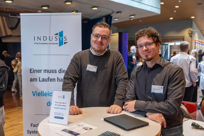 Zwei Männer mit Namensschildern sitzen an einem Tisch vor einem Banner der Firma INDUSYS.