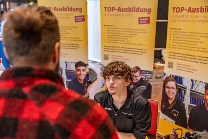 Ein junger Mann im DHL-Shirt unterhält sich mit einer Person im Vordergrund vor gelben Plakaten für Ausbildungen.