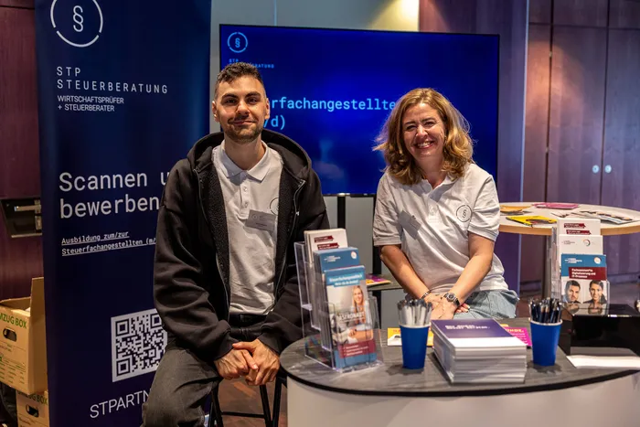 Ein Mann und eine Frau sitzen lächelnd an einem Infostand der STP Steuerberatung mit Broschüren und Werbematerialien.
