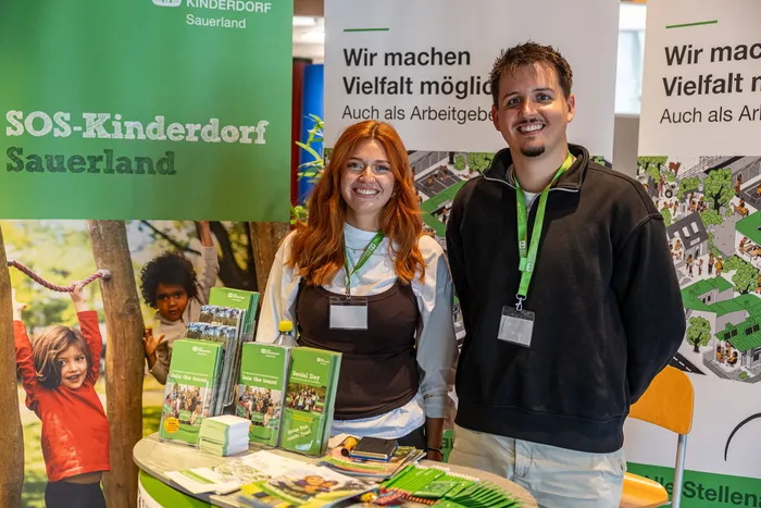 Zwei lächelnde Mitarbeiter stehen an einem Infostand von SOS-Kinderdorf Sauerland mit verschiedenen Broschüren auf dem Tisch.