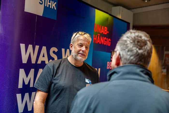 Ein grauhaariger Mann im schwarzen T-Shirt unterhält sich mit einer anderen Person vor einem blauen Banner mit dem Schriftzug „UNABHÄNGIG“ und dem SIHK-Logo.