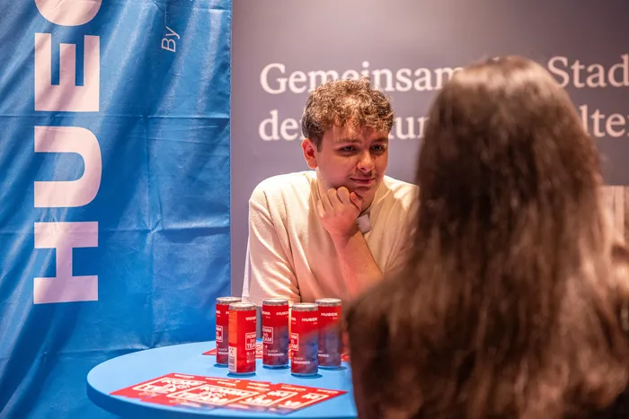 Ein junger Mann blickt aufmerksam zu einer Person mit langen dunklen Haaren, die ihm gegenüber an einem blauen Tisch mit Getränkedosen sitzt.