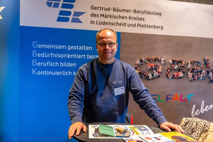 Ein lächelnder Mann mit Brille und blauem Sweatshirt steht vor einem Banner des Gertrud-Bäumer-Berufskollegs.