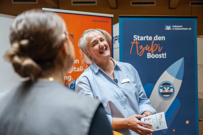 Eine lachende Frau in einem hellblauen Hemd steht an einem Messestand vor einem blauen Banner mit der Aufschrift „Starte den Azubi Boost!“.