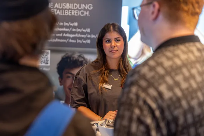 Eine junge Frau steht an einem Informationsstand vor einem Plakat über Ausbildungen im Metallbereich und unterhält sich mit Personen im Vordergrund.