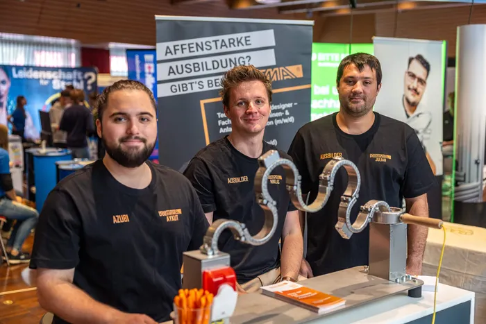Drei Männer, ein Auszubildender und zwei Ausbilder, präsentieren ein geschwungenes Metallobjekt an einem Messestand.