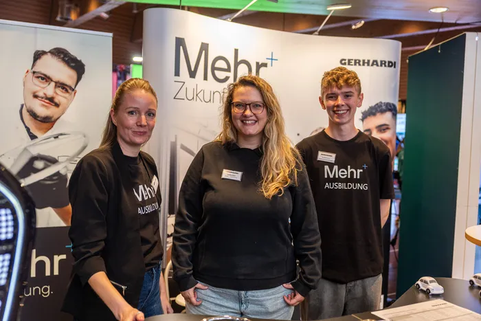 Zwei Frauen und ein junger Mann lächeln vor einem Messestand mit dem Schriftzug „Mehr+ Ausbildung“ der Firma Gerhardi.