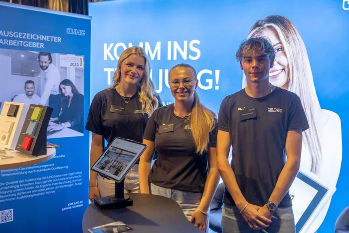 Drei junge Menschen in dunkelblauen JUNG-T-Shirts stehen vor einem blauen Banner mit der Aufschrift „KOMM INS TEAM JUNG!“.