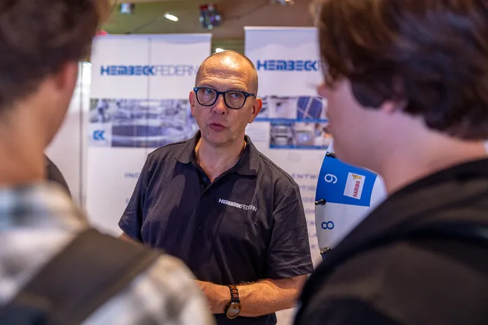 Ein Mann mit Brille und einem Hembeck-Federn-Polohemd im Gespräch mit Besuchern an einem Messestand.