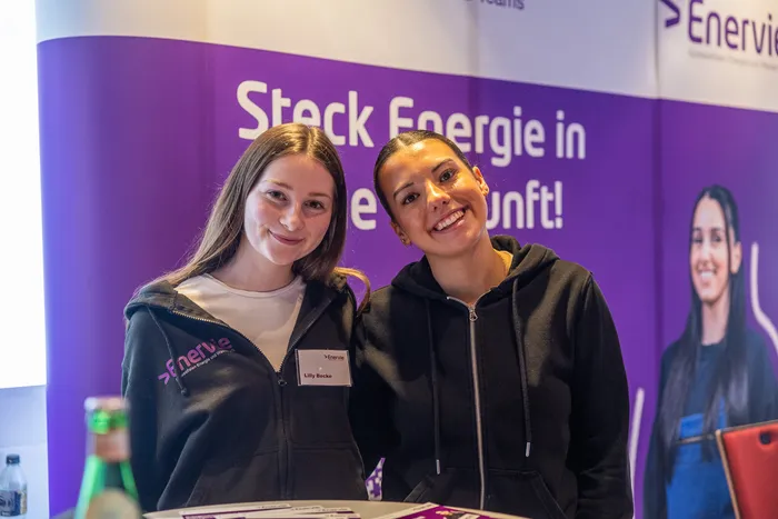 Zwei junge Frauen lächeln vor einem lila Banner mit der Aufschrift „Steck Energie in die Zukunft!“ und dem EnerVie-Logo.