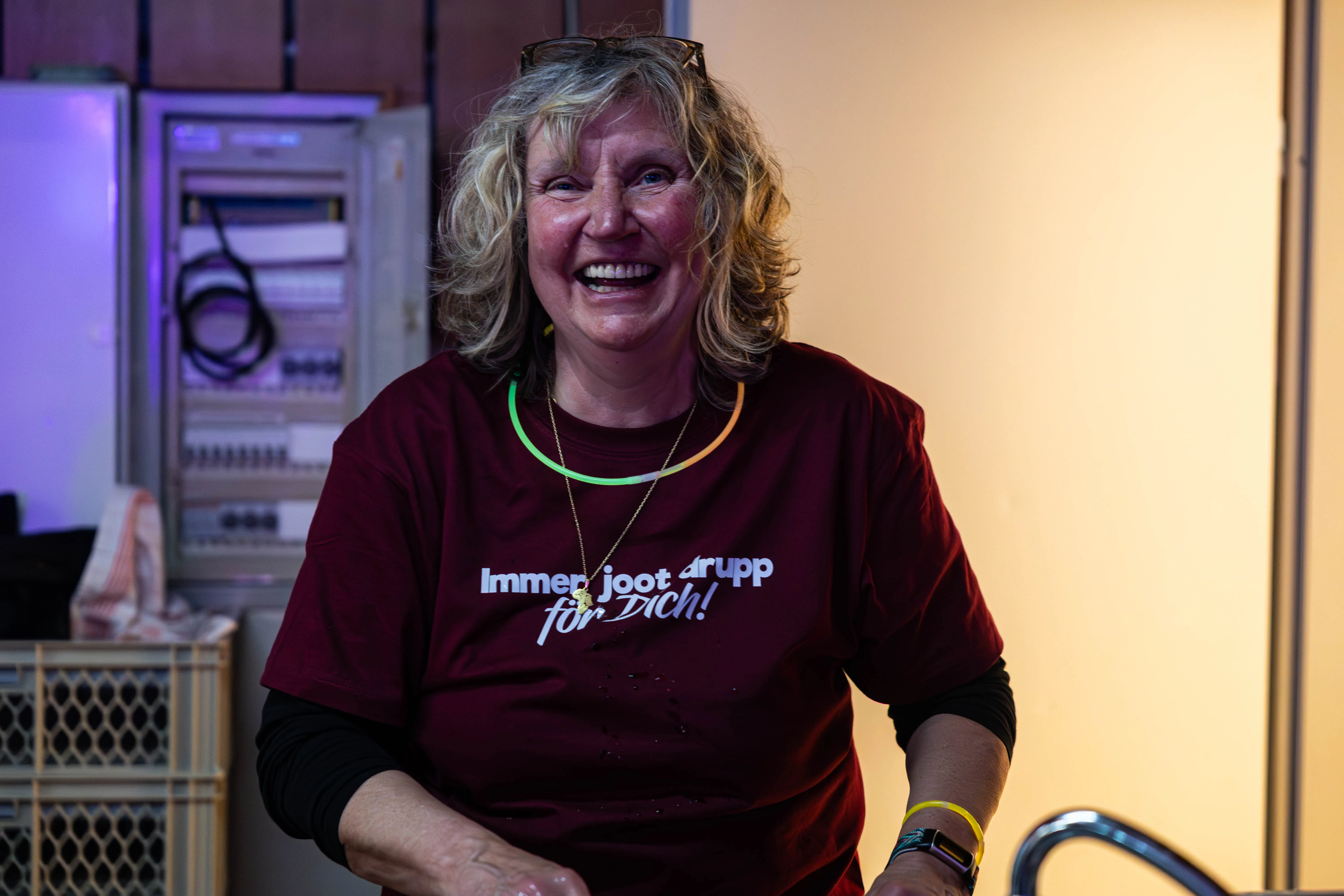 Lachende Frau mit blonden Locken und rotem T-Shirt mit Aufschrift „Immer joot drupp für Dich!“. Im Hintergrund sind technische Geräte und ein schwarzer Kasten zu sehen.
