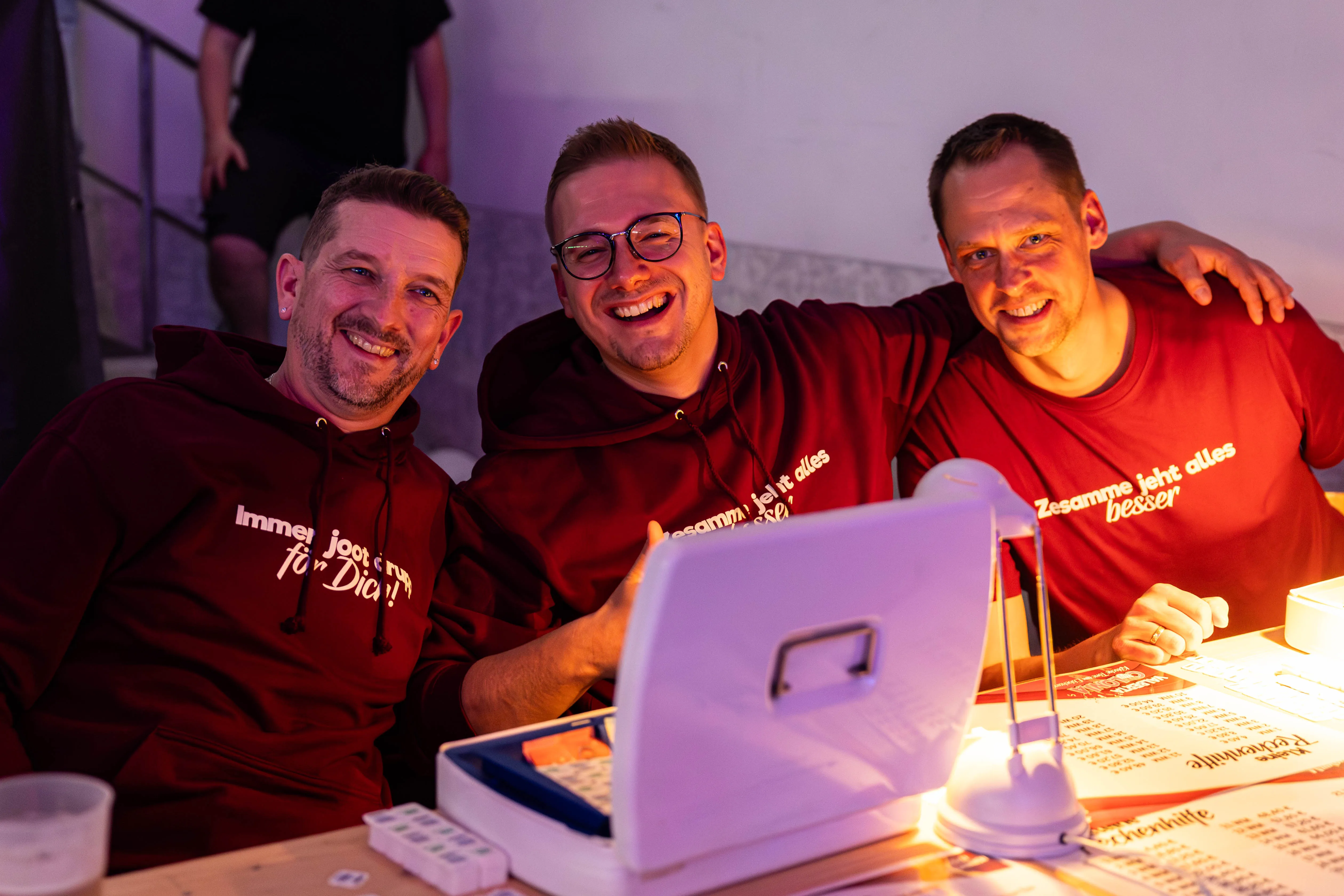Drei lächelnde Männer in roten Hoodies mit Aufschriften stehen an einem Tisch mit Getränken und Notizblöcken. Im Hintergrund sind weitere Personen und eine Bühne zu erkennen.