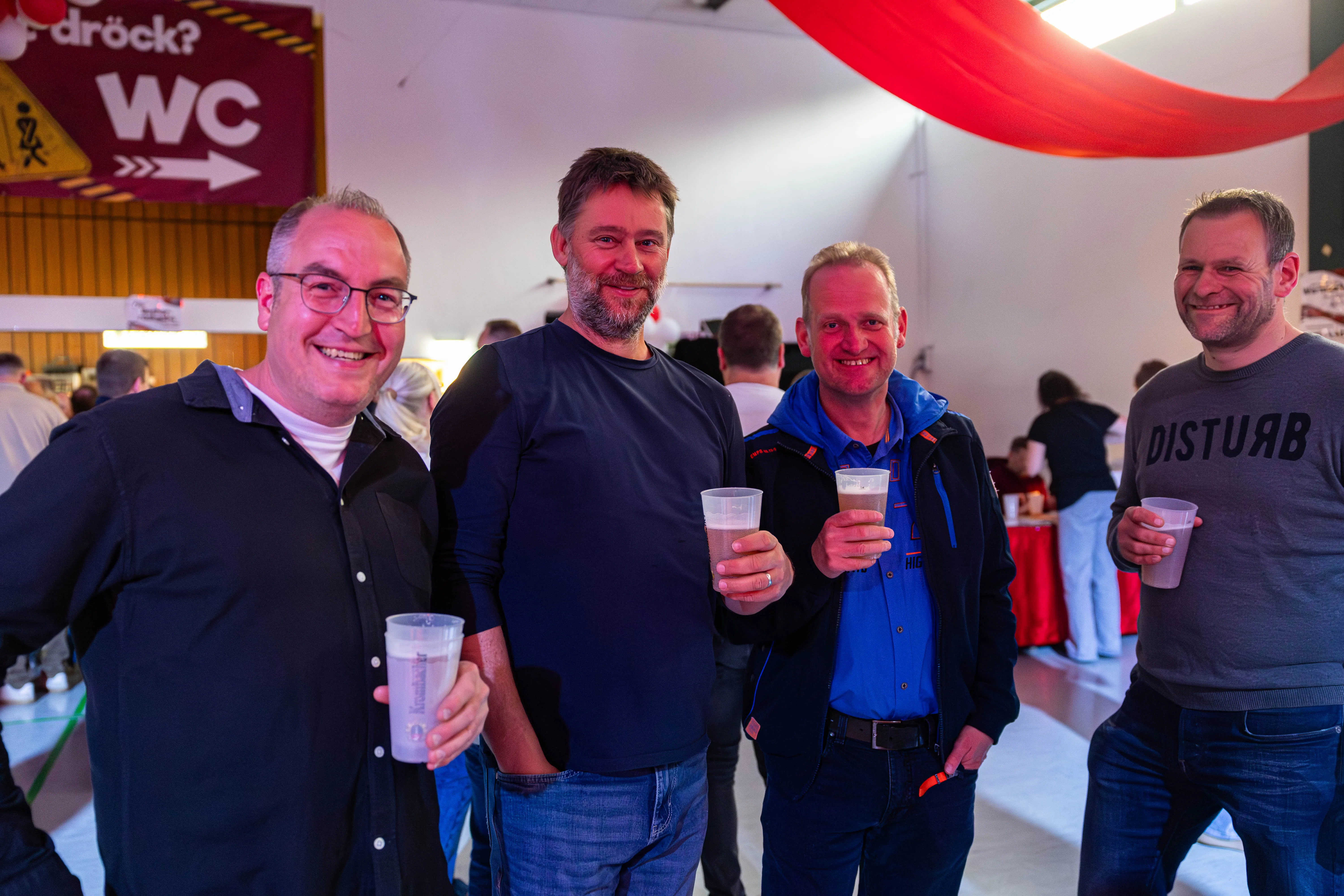 Vier lächelnde Männer stehen eng zusammen und halten jeweils ein Glas in der Hand, im Hintergrund ein Festivalzelt mit dem Hinweis auf die Toiletten.
