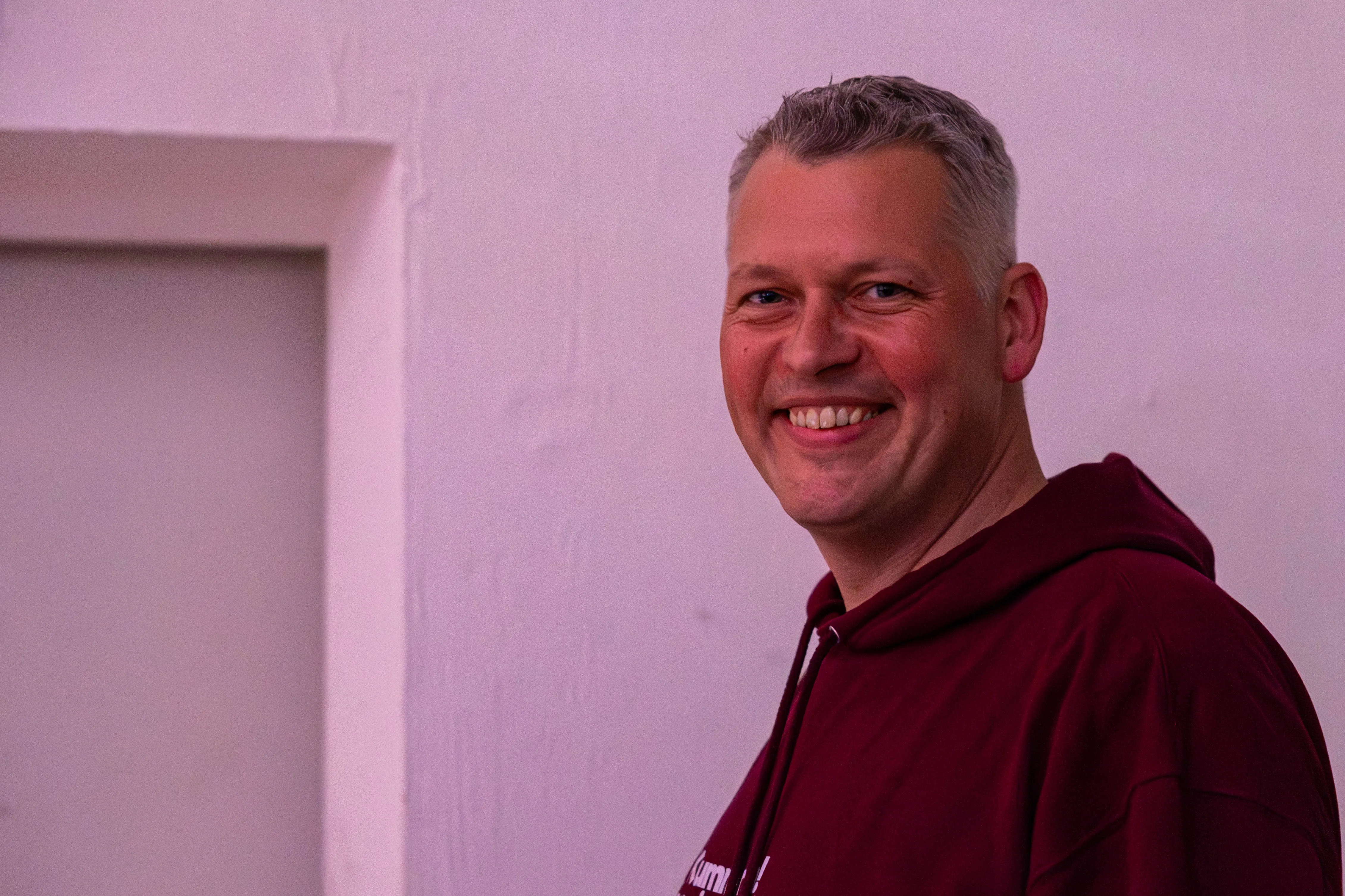 Lachender Mann mit kurz geschnittenem, grau meliertem Haar und rotem Kapuzenpullover vor pinkfarbener Wand.