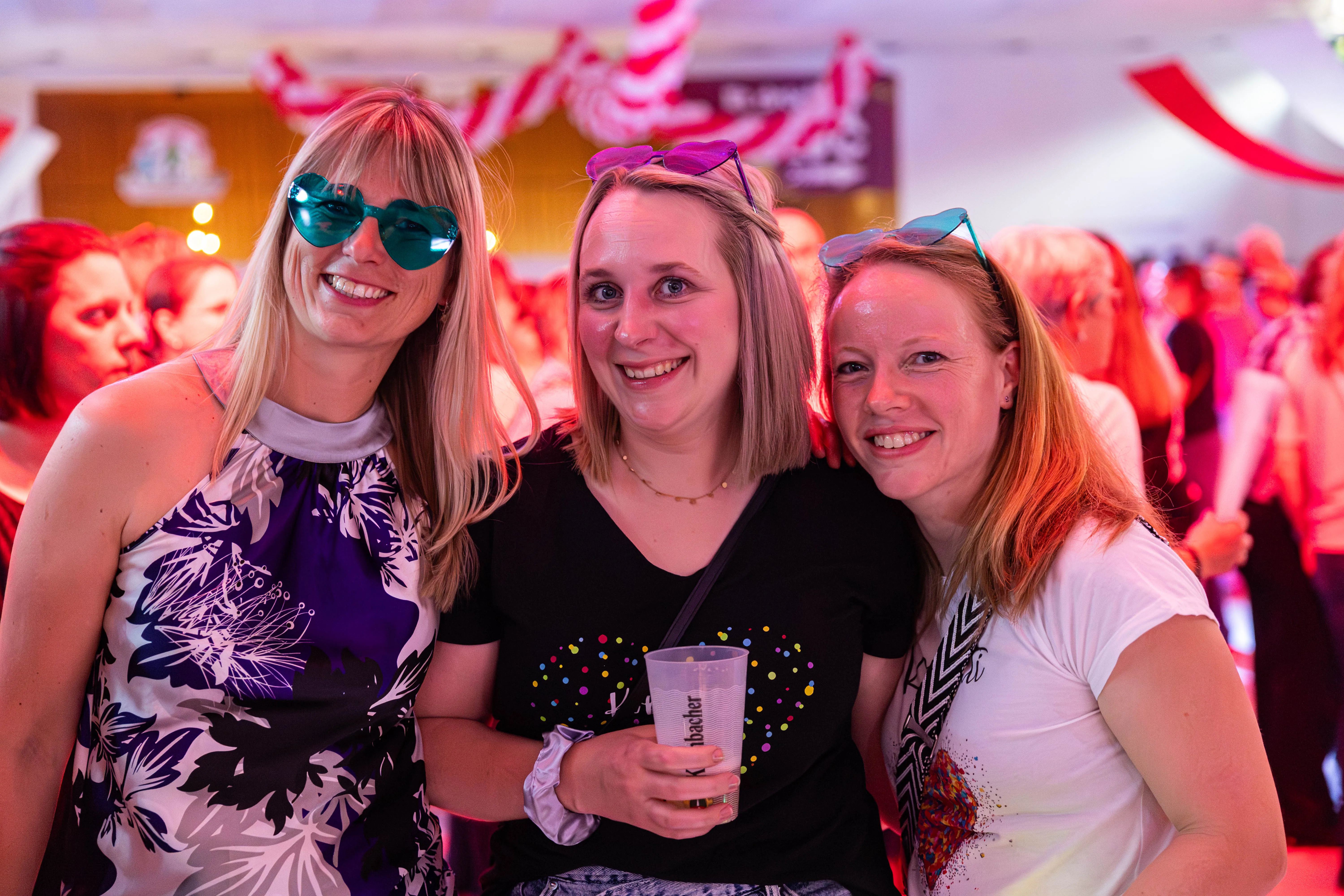 Drei lächelnde Frauen posieren eng nebeneinander, jede mit Partyaccessoires wie Brillen und Haarreifen. Im Hintergrund sind weitere Menschen und eine festliche Beleuchtung zu sehen.
