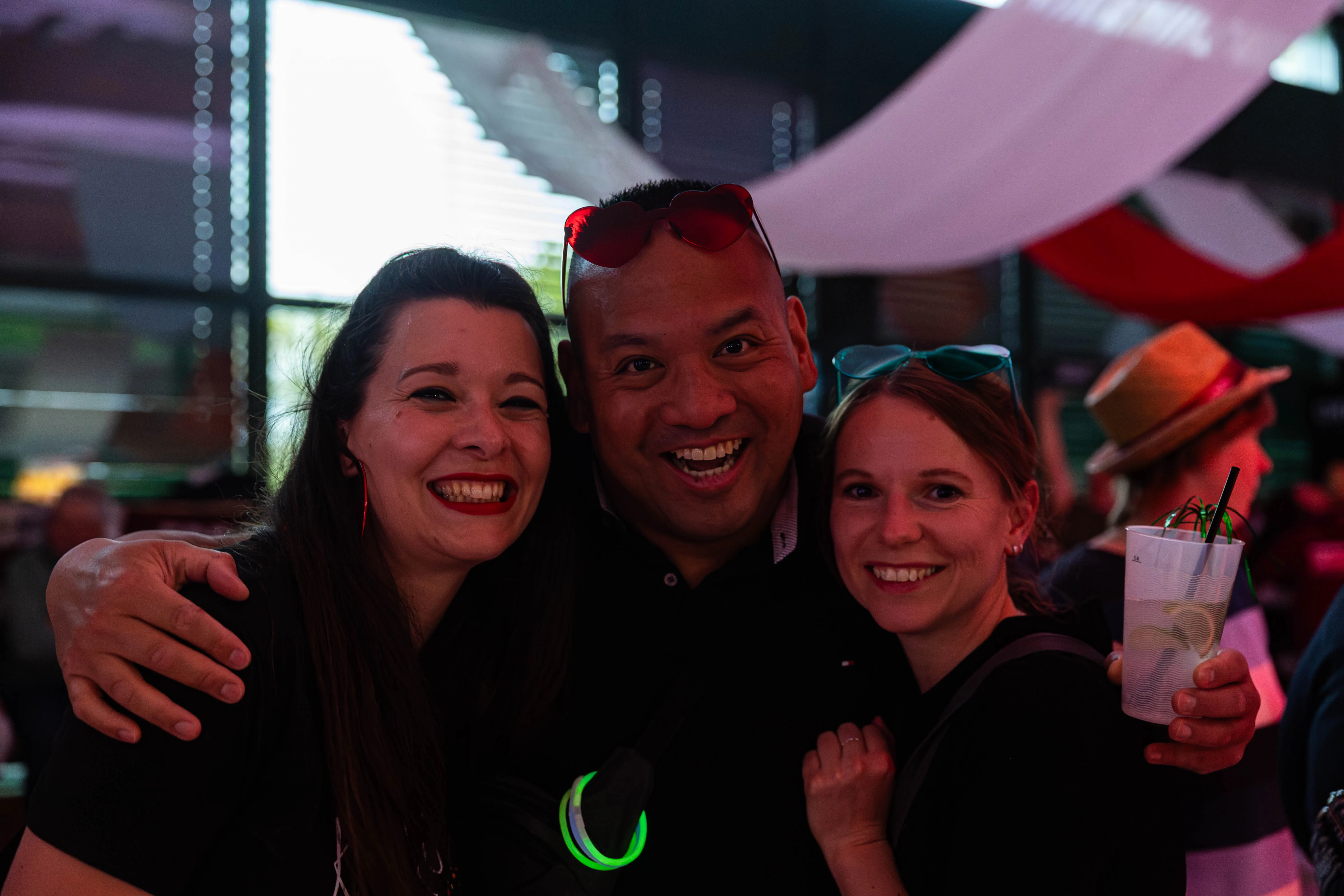 Drei lächelnde Menschen posieren eng umschlungen für ein Foto. Im Hintergrund sind weitere Festivalbesucher und Bühnenelemente zu erkennen.