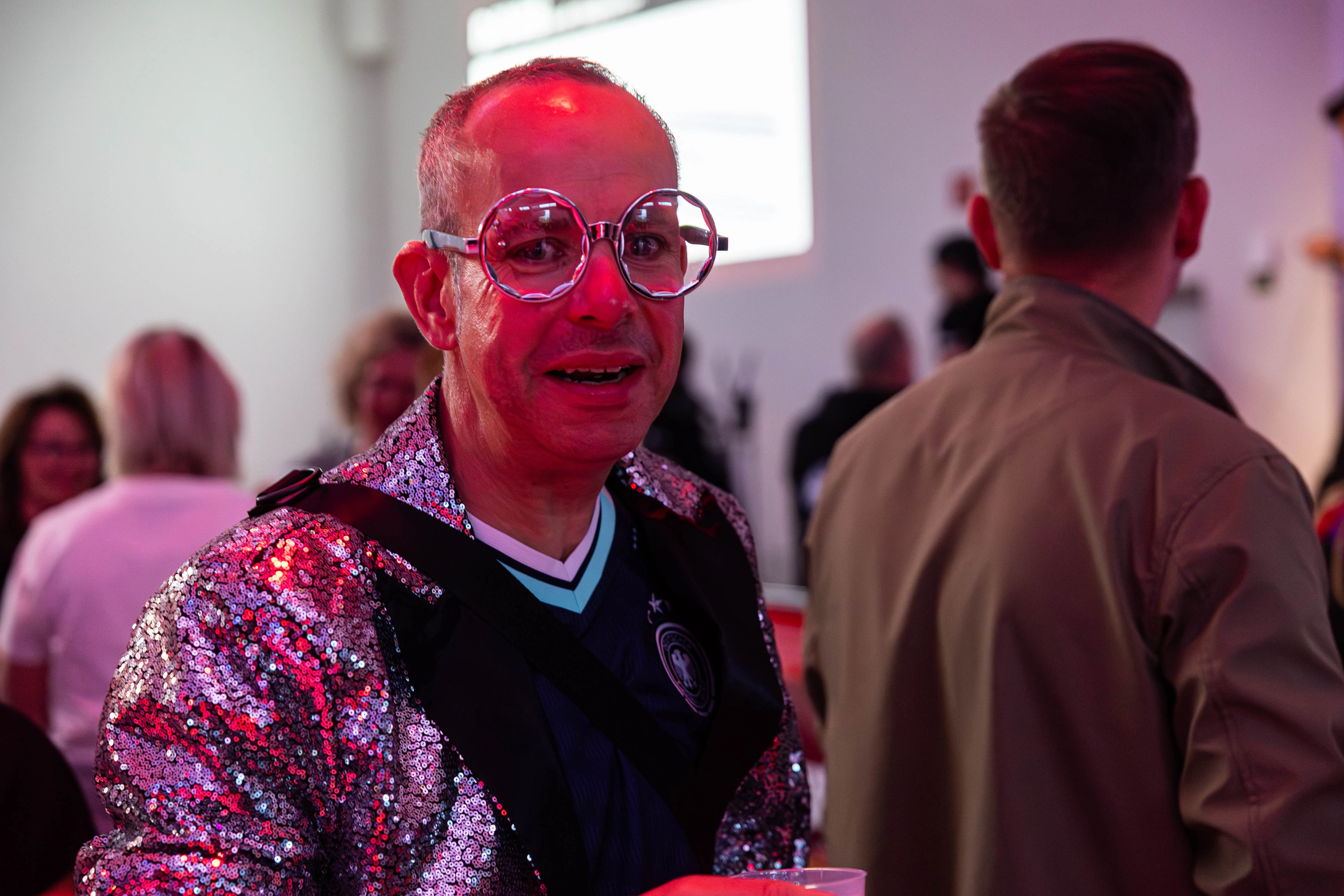 Mann mit großer Brille und glitzernder Weste unterhält sich auf einem Festival mit einem anderen Mann im Hintergrund.