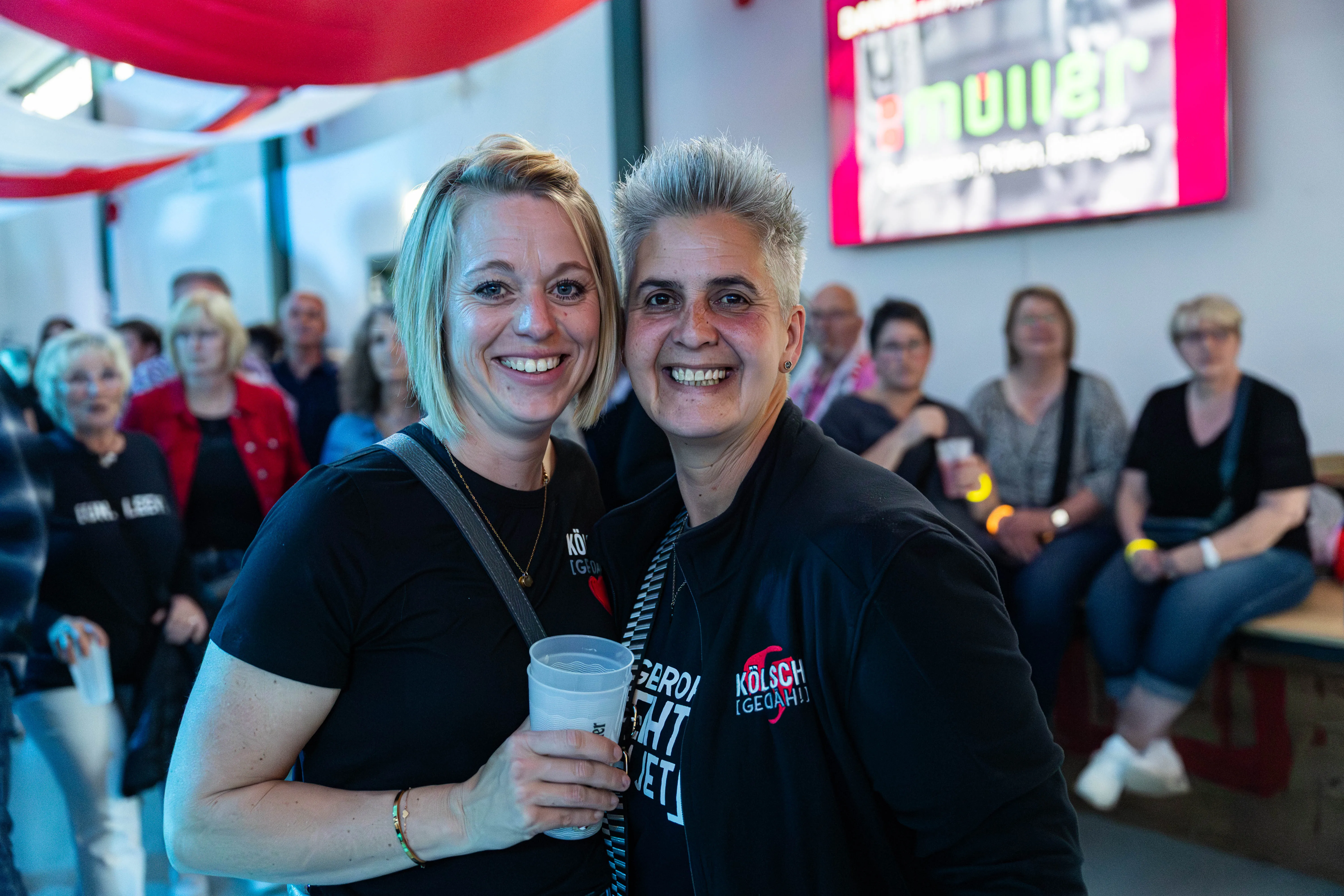 Zwei lächelnde Frauen im Vordergrund, die Kölsch-T-Shirts tragen und Gläser halten, umgeben von Menschen auf einem Festivalgelände.