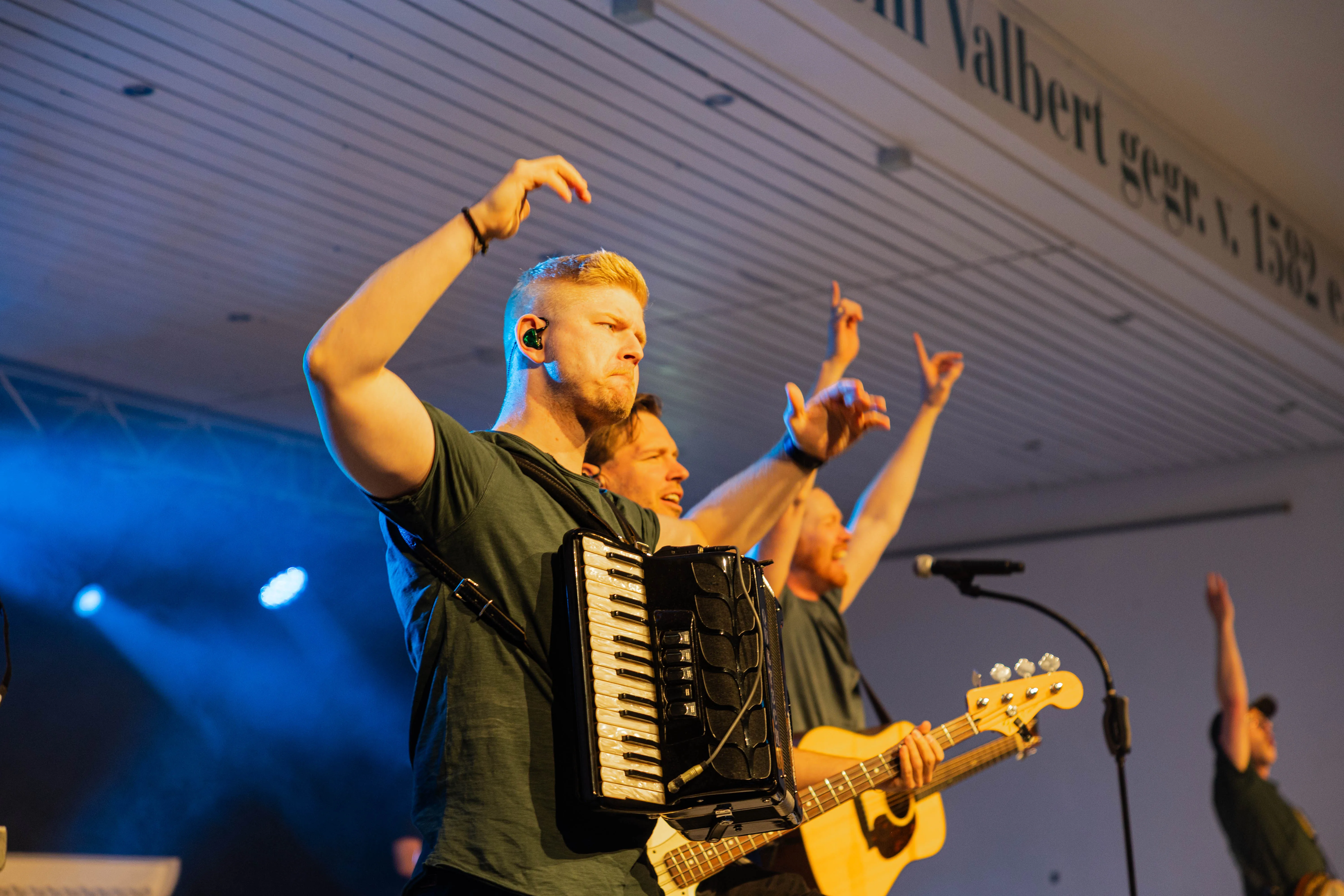 Ein Musiker spielt auf einem Akkordeon, während zwei weitere Bandmitglieder auf einer Bühne singen und ein Bassist im Hintergrund steht.