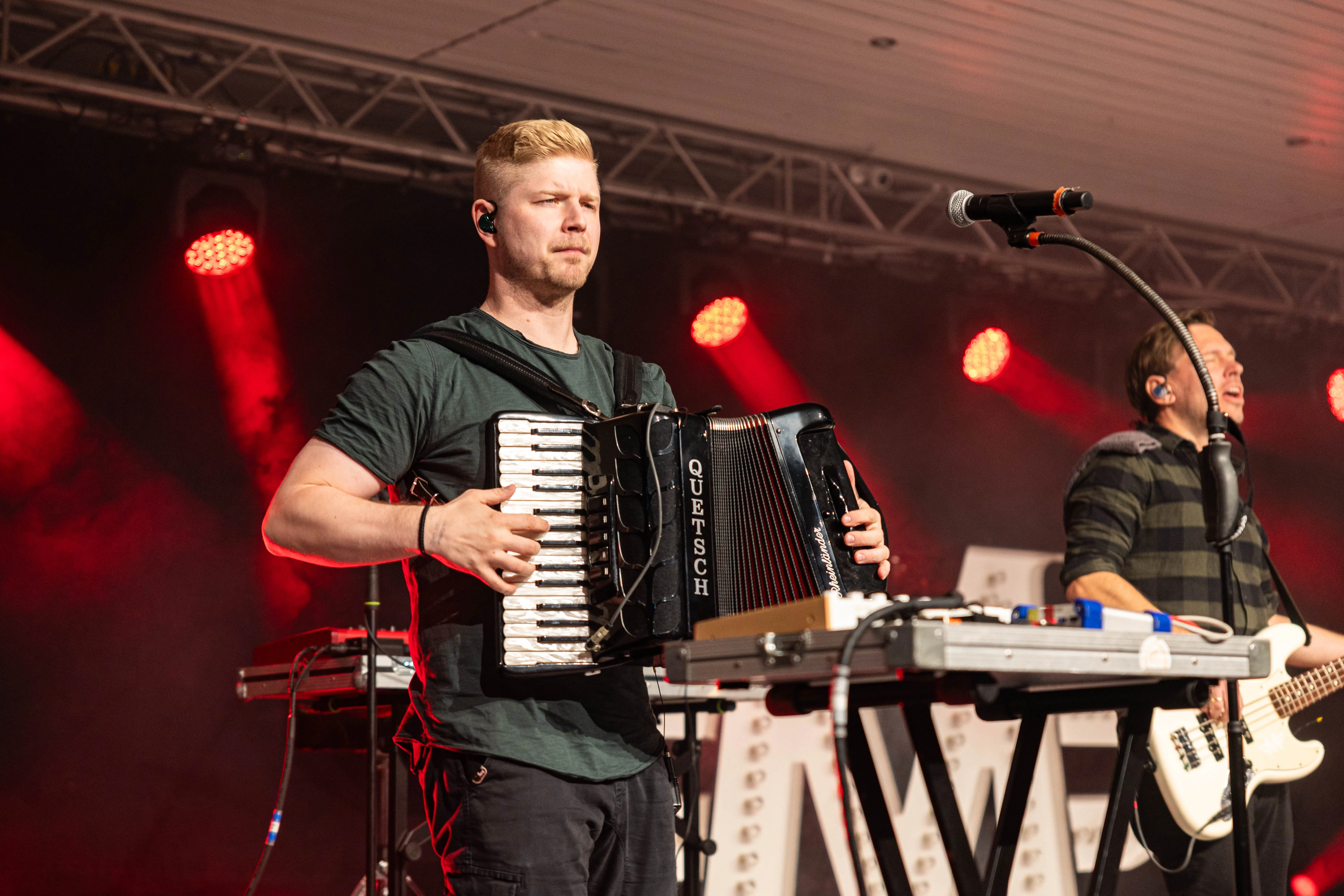 Ein Musiker spielt auf einem Akkordeon vor Publikum, während ein zweiter Musiker im Hintergrund ein Bassinstrument bedient. Im Hintergrund leuchten rote Bühnenlichter.