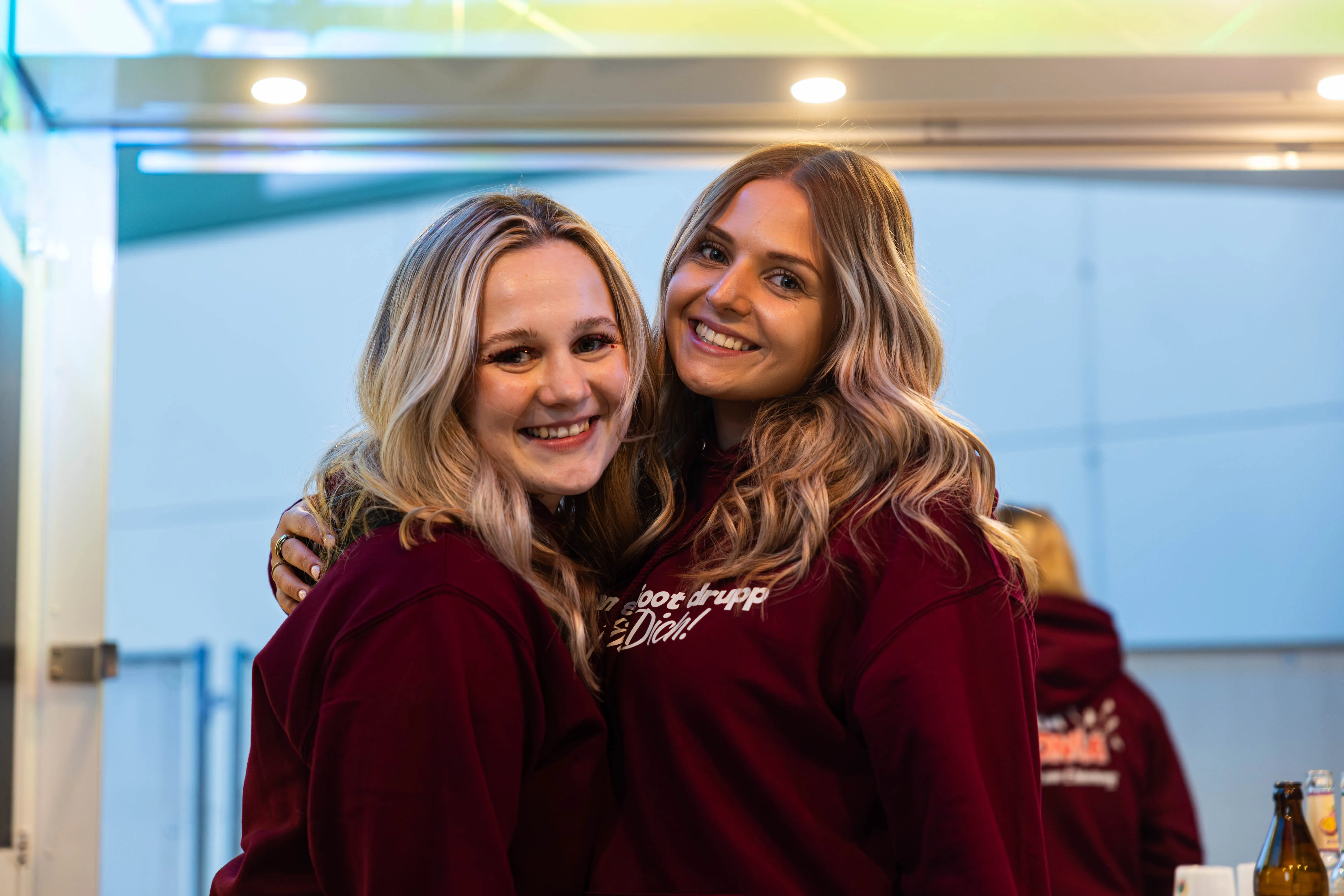 Zwei junge Frauen mit blonden Haaren umarmen sich lachend und tragen dunkelrote Pullover mit Aufschrift. Im Hintergrund sind weitere Personen und ein heller Raum zu sehen.