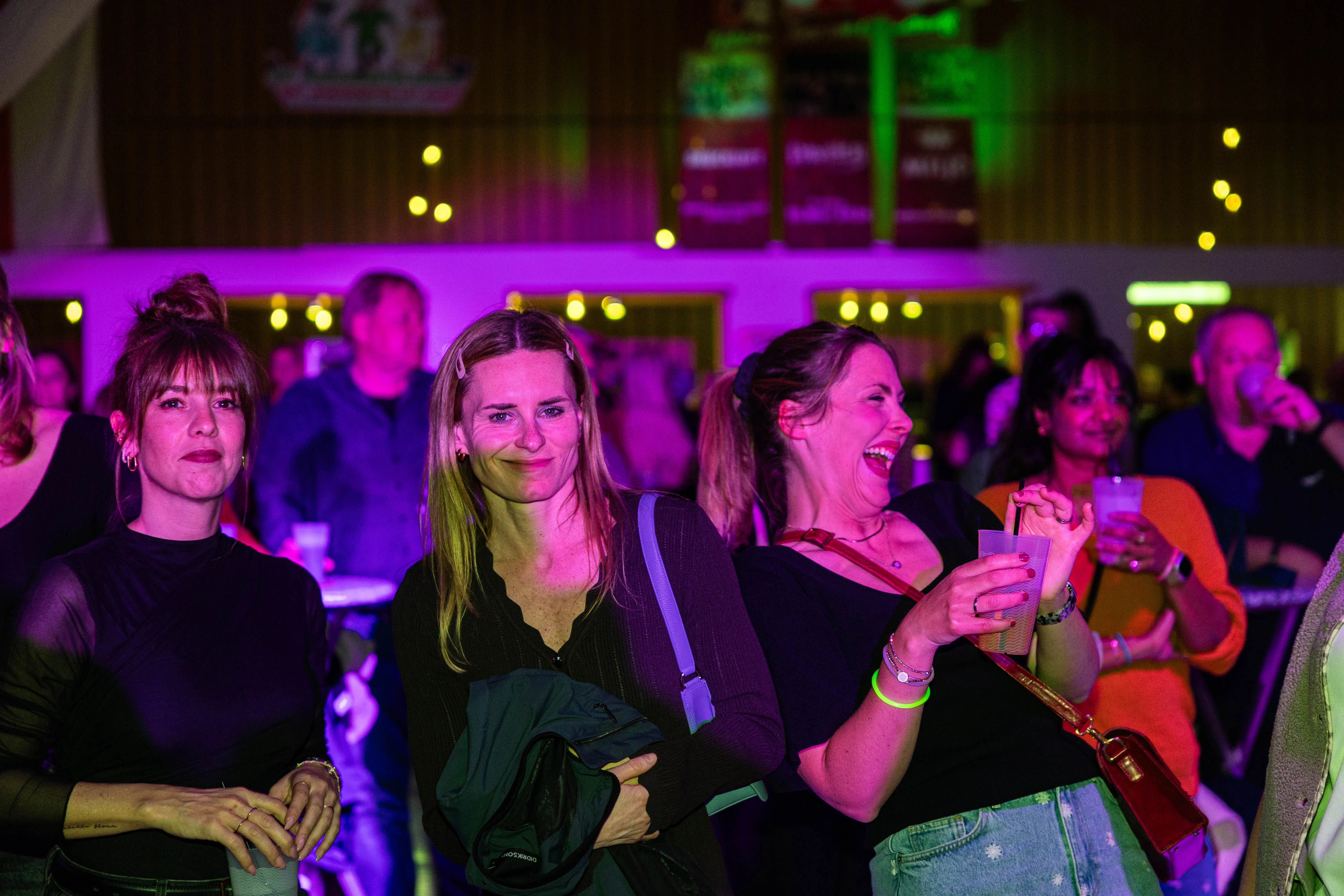 Drei lächelnde Frauen im Vordergrund eines Konzerts, im Hintergrund weitere Konzertbesucher und eine Bühne mit bunten Lichtern.