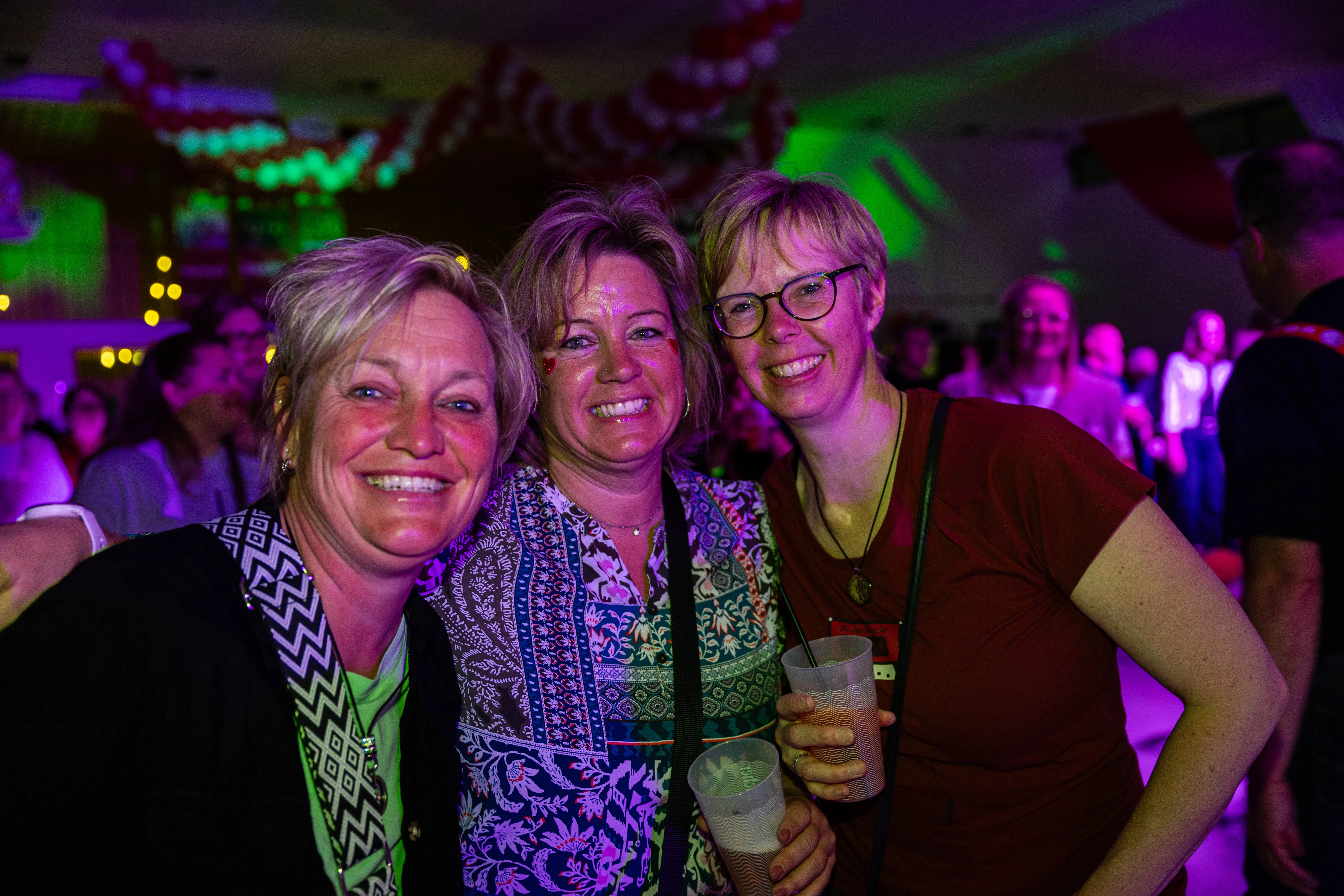 Drei lächelnde Frauen posieren eng zusammen vor einer Menschenmenge auf einem Konzert. Im Hintergrund sind bunte Lichter und Dekorationen zu sehen.