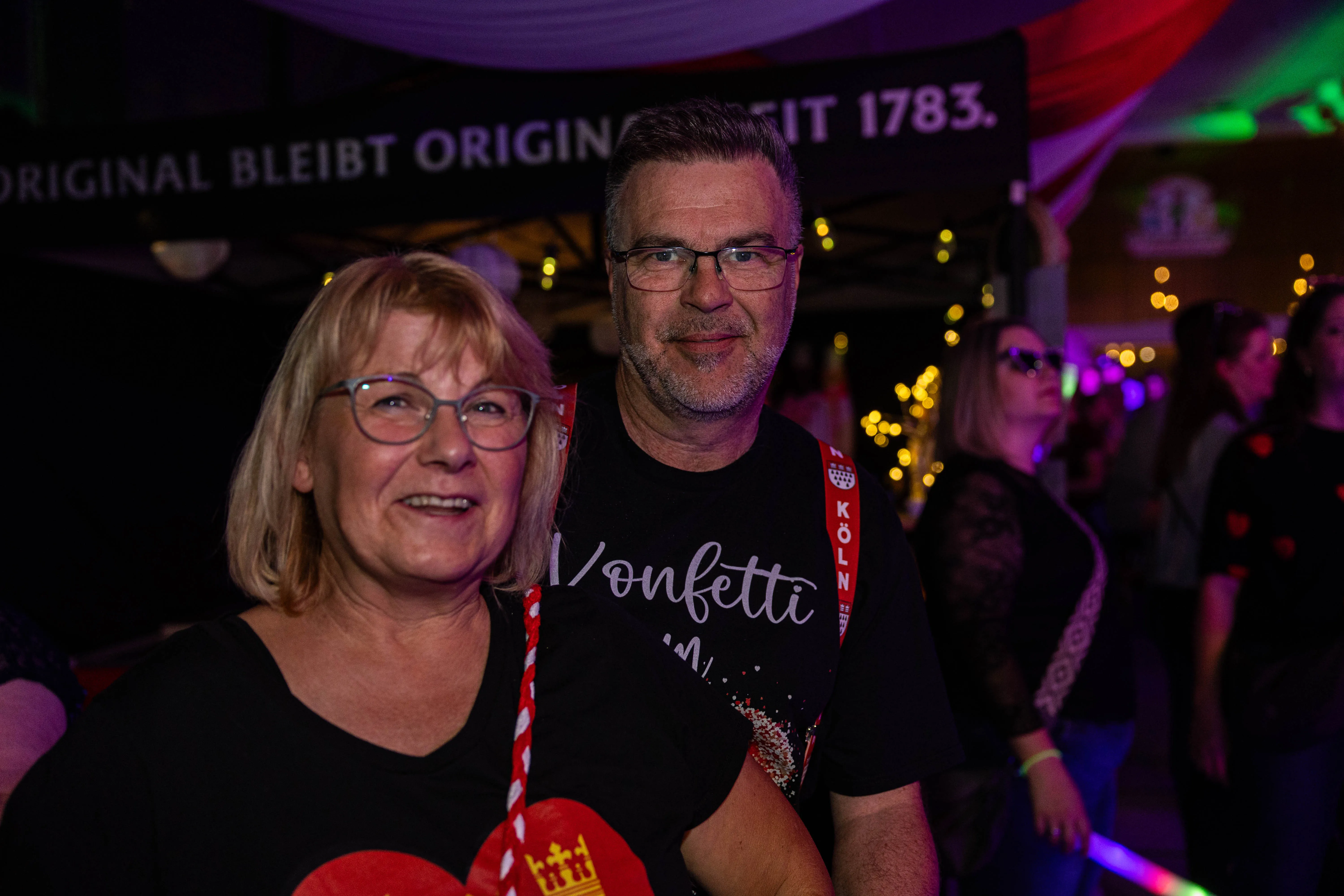 Zwei lächelnde Menschen, eine Frau und ein Mann, vor einer Menschenmenge und einer dunklen Bühne mit Lichtern und Bannern. Der Mann trägt ein T-Shirt mit der Aufschrift „Konfetti“ und eine rote Schärpe mit der Aufschrift „Köln“.