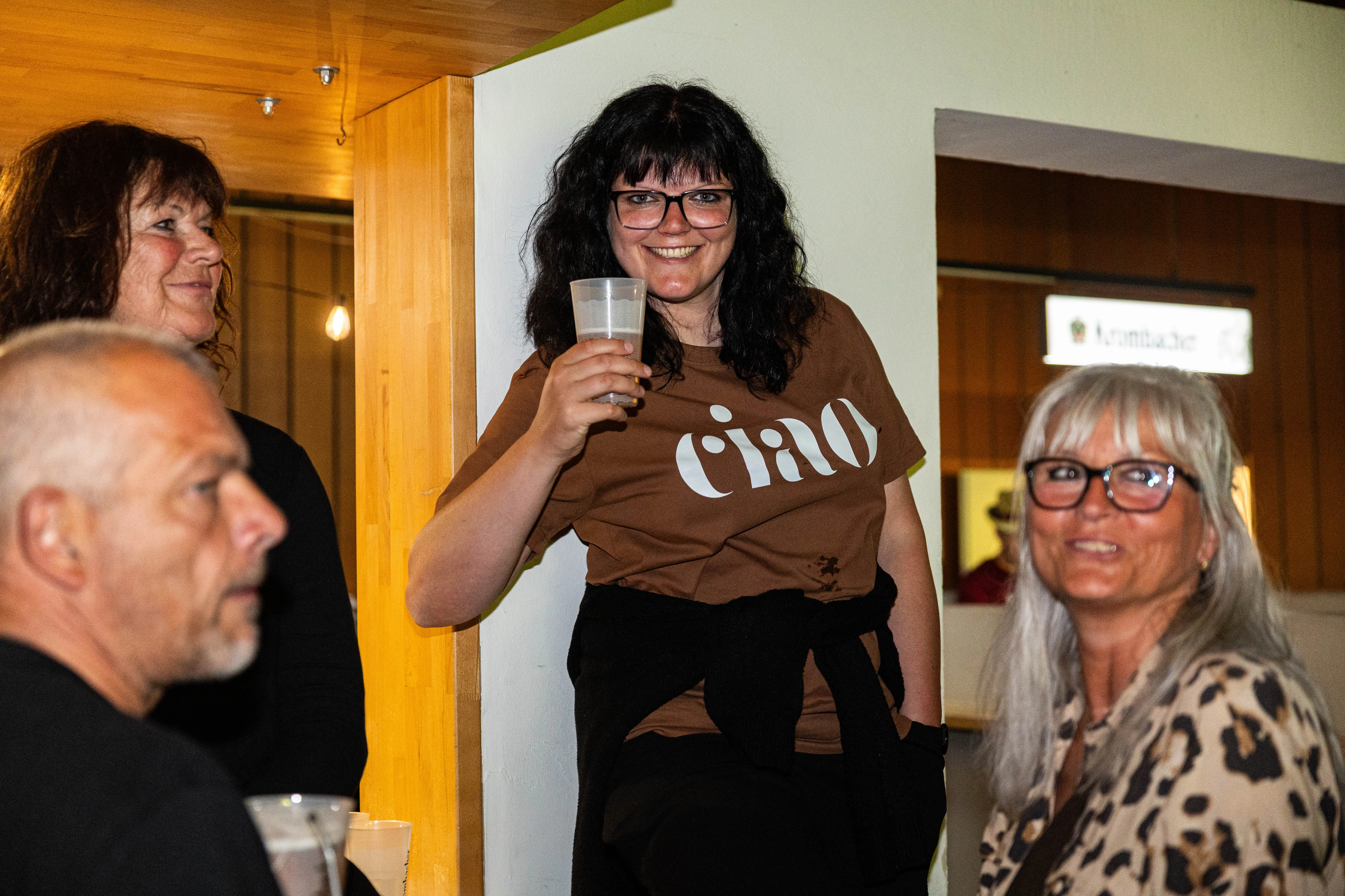 Eine Frau mit dunklen Haaren und Brille lächelt in die Kamera, während sie ein Glas hält. Im Hintergrund sind weitere Personen und ein Holzvertäfelung zu sehen.