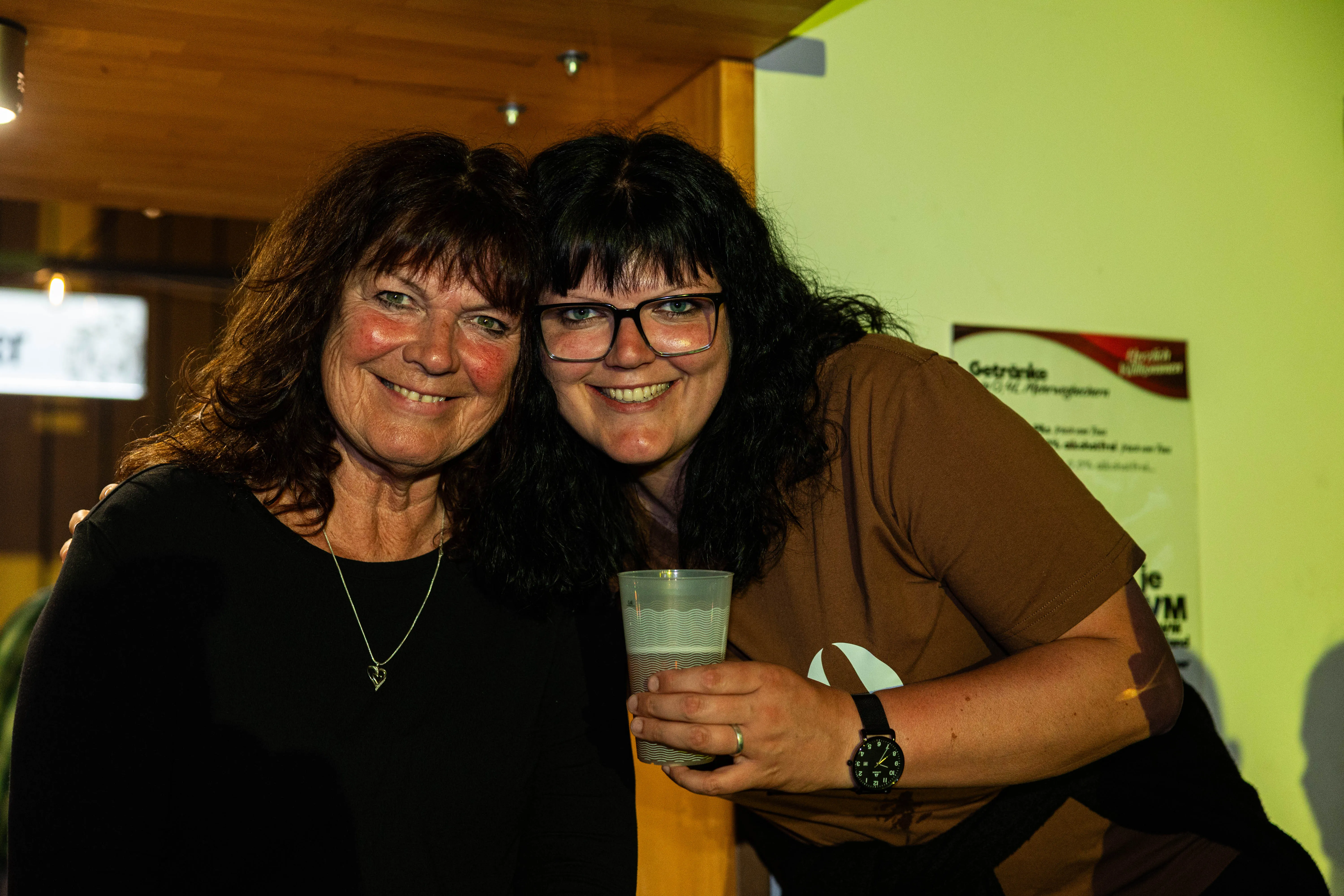 Zwei lächelnde Frauen umarmen sich vor einem hellen Hintergrund. Die Frau rechts hält ein Glas mit Flüssigkeit und trägt eine Smartwatch.