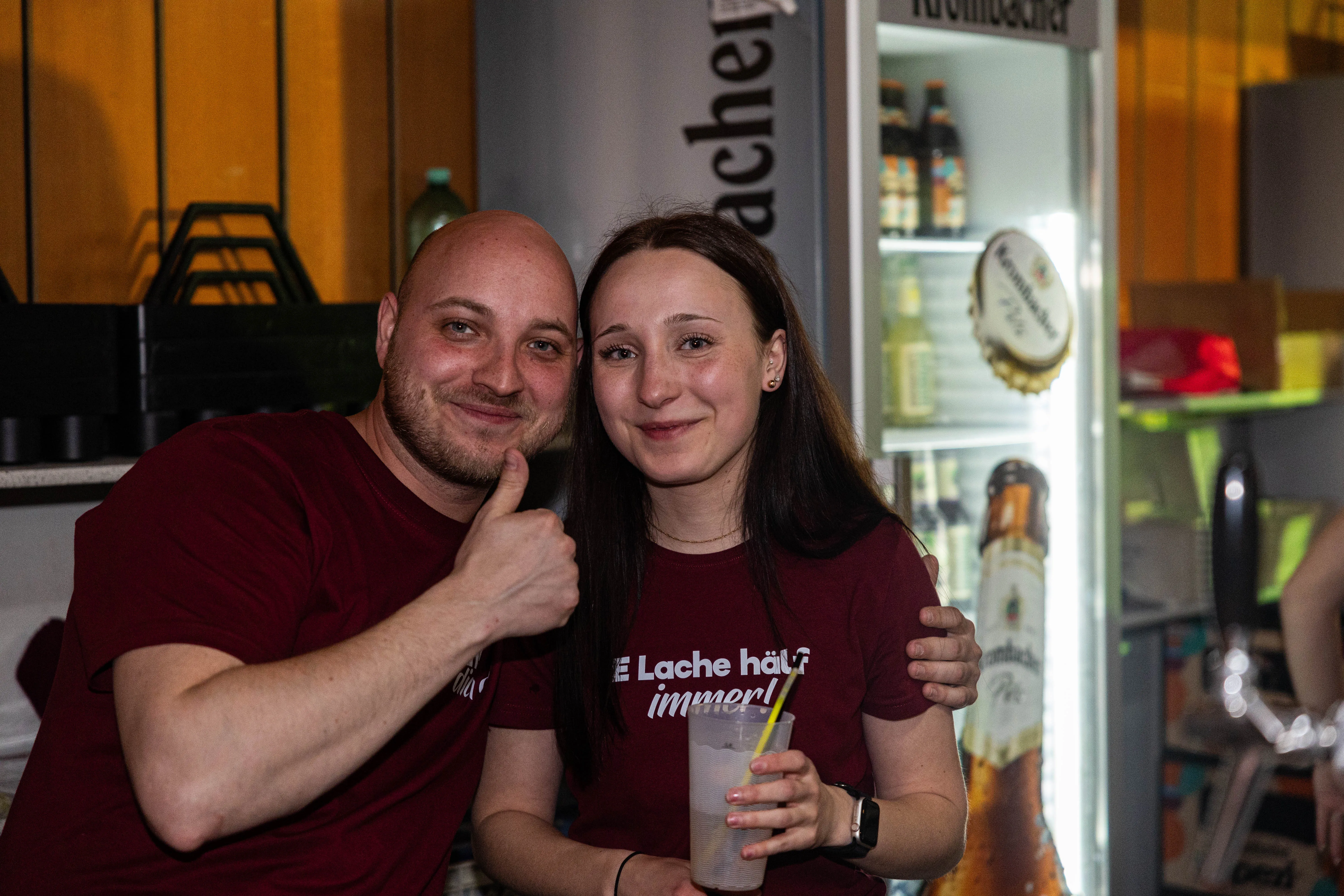 Zwei lächelnde Menschen, ein Mann und eine Frau, stehen vor einer Bar mit Getränken. Die Frau hält ein Glas, der Mann zeigt Daumen hoch.