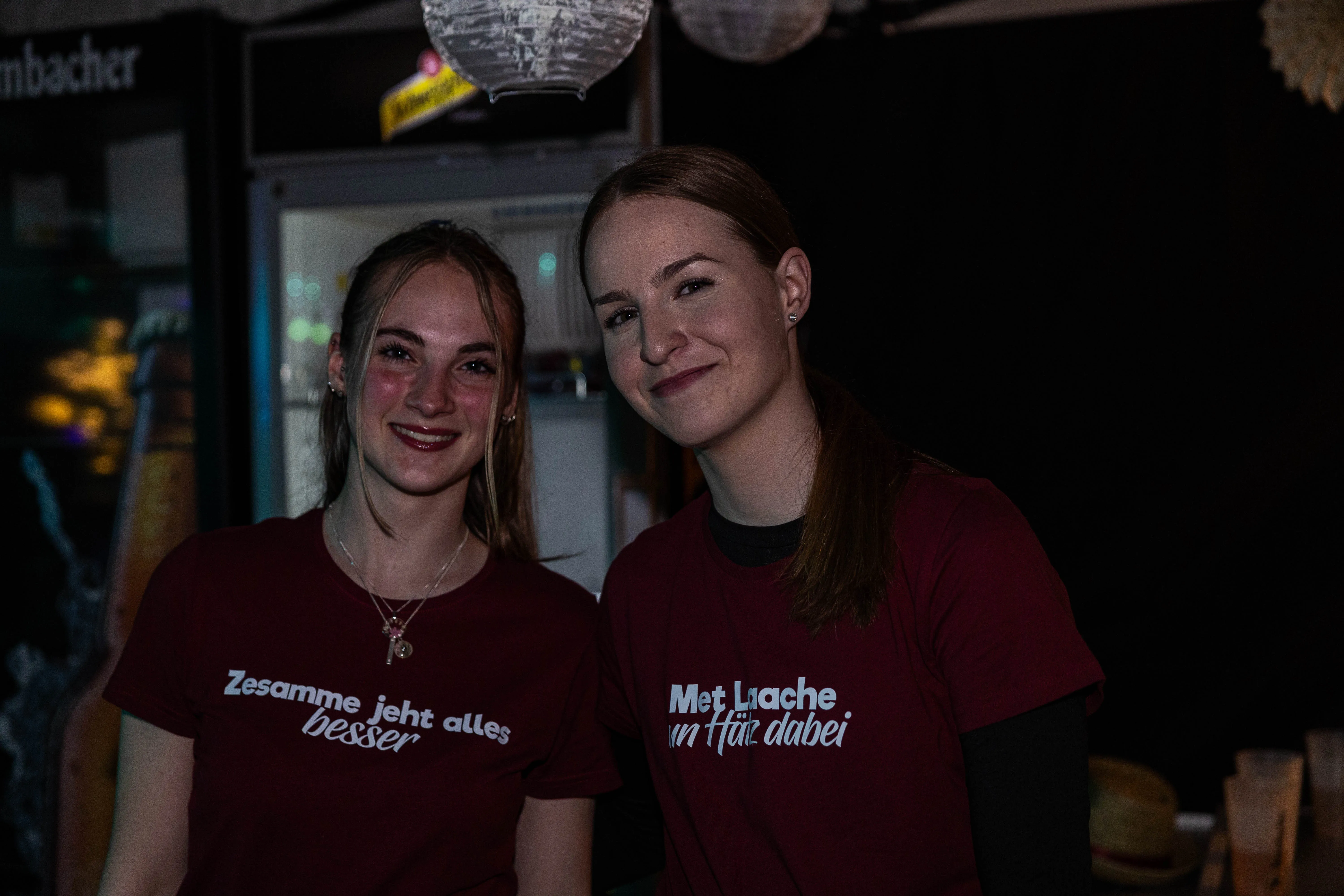 Zwei junge Frauen mit roten T-Shirts und Aufschriften lächeln in die Kamera. Im Hintergrund ist eine Bar mit Getränken zu sehen.