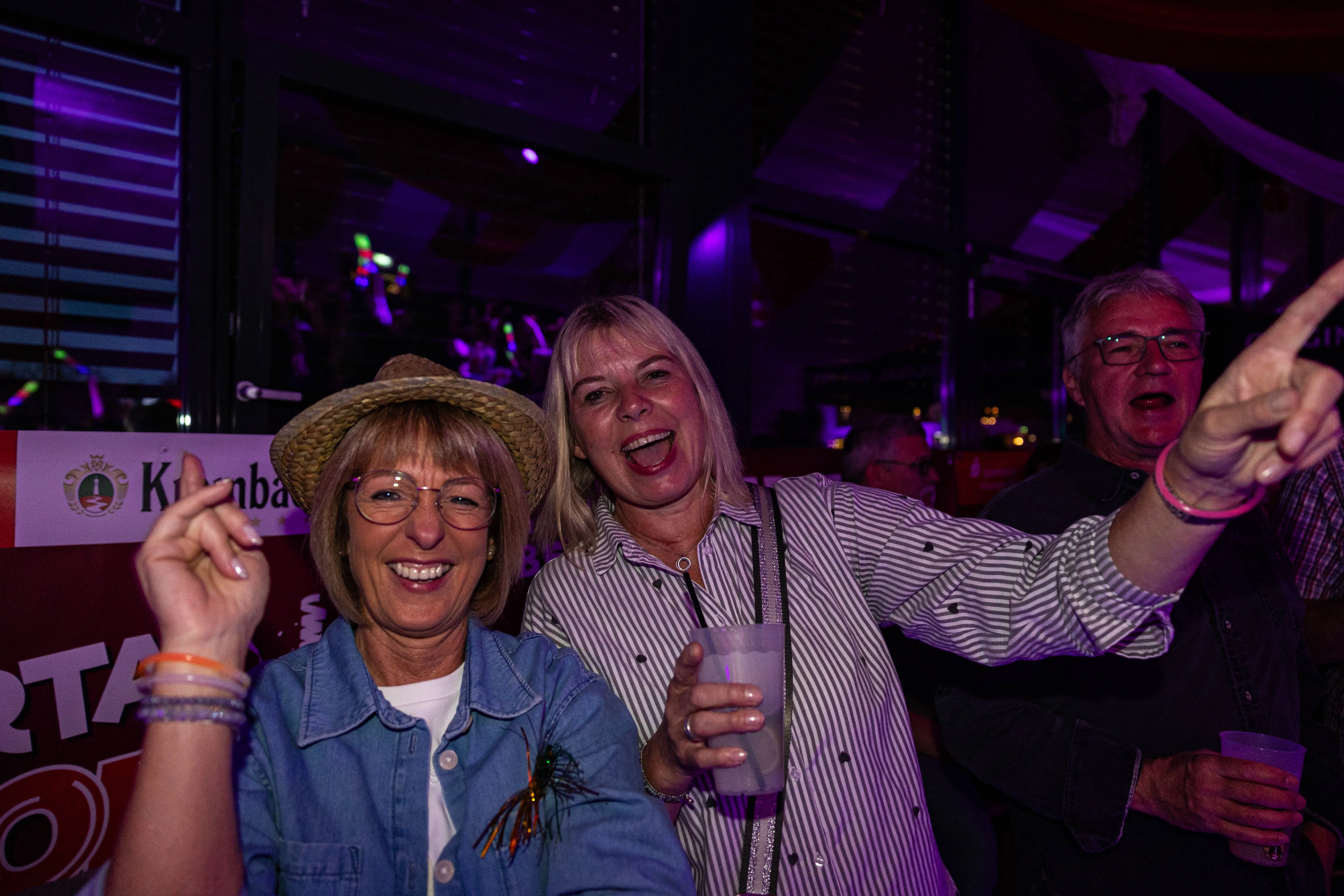 Drei ausgelassene Menschen feiern auf dem Kölschmusikfestival „Valberta Colonia“, eine Frau mit Strohhut und zwei Männer mit erhobenen Händen und Gläsern.