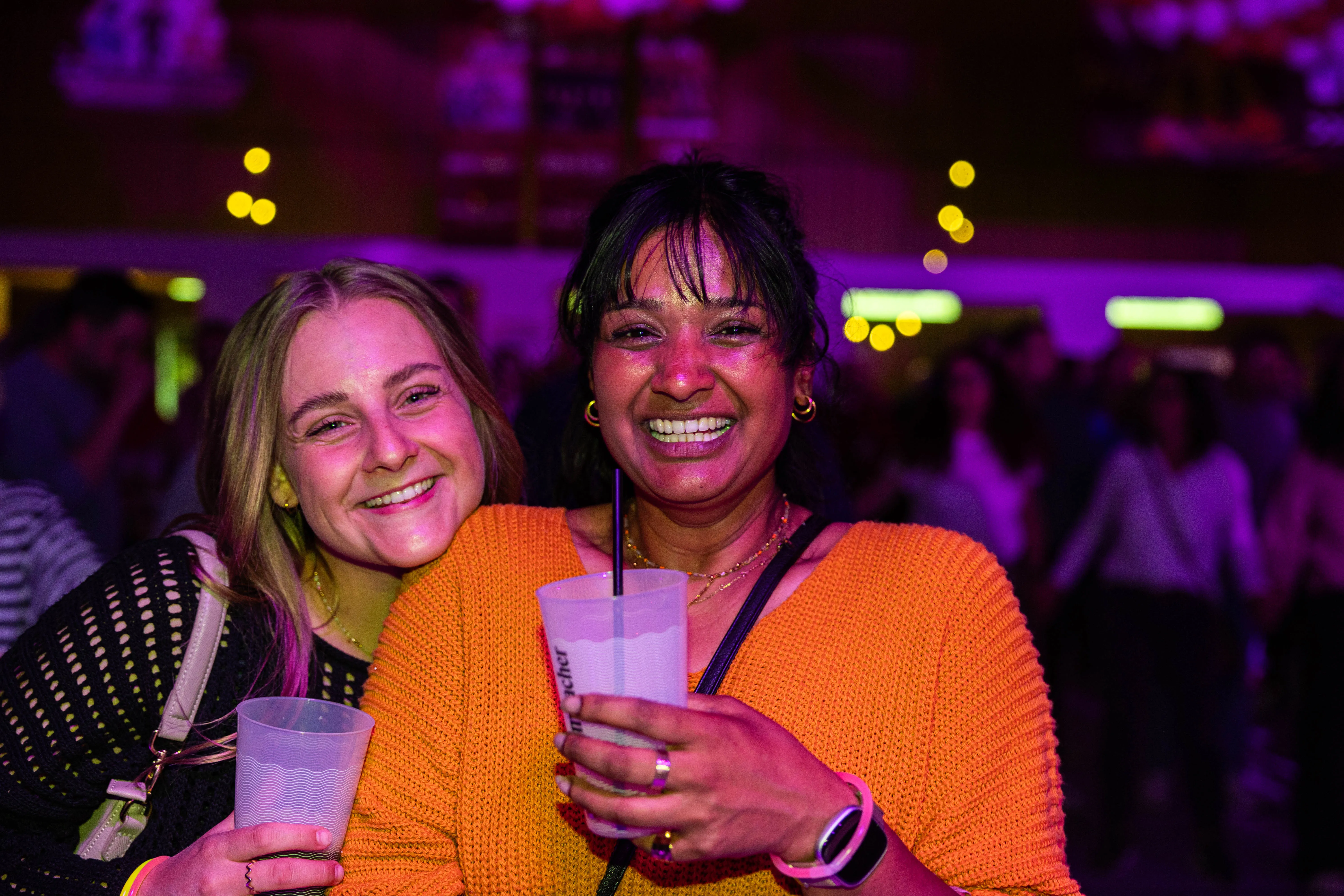 Zwei lächelnde Frauen vor einer Menschenmenge bei einem Konzert. Die Frau rechts hält ein Getränk mit Strohhalm.