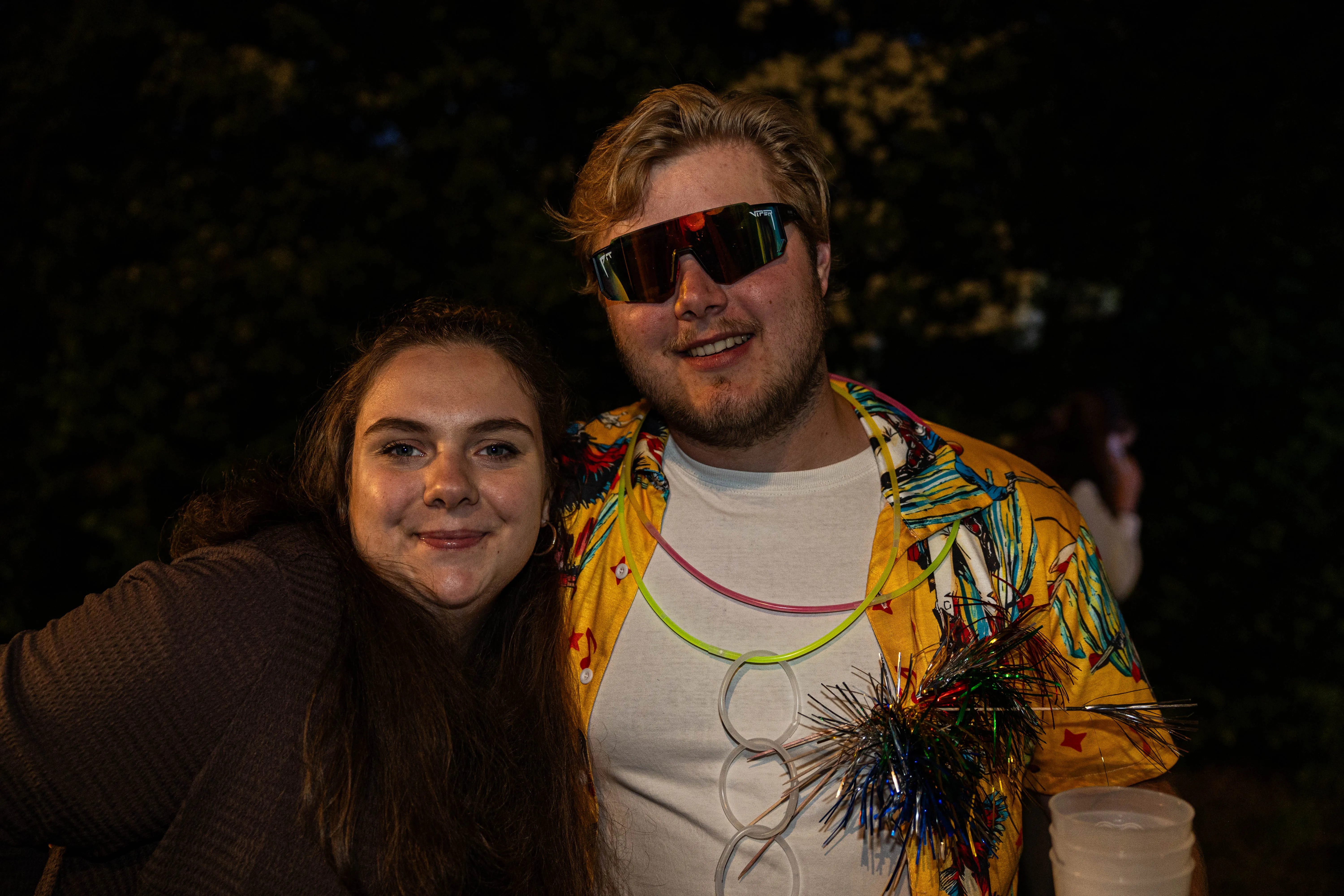 Zwei junge Erwachsene lächeln in die Kamera. Der Mann trägt eine bunte Jacke und Sonnenbrille, die Frau hat lange braune Haare.