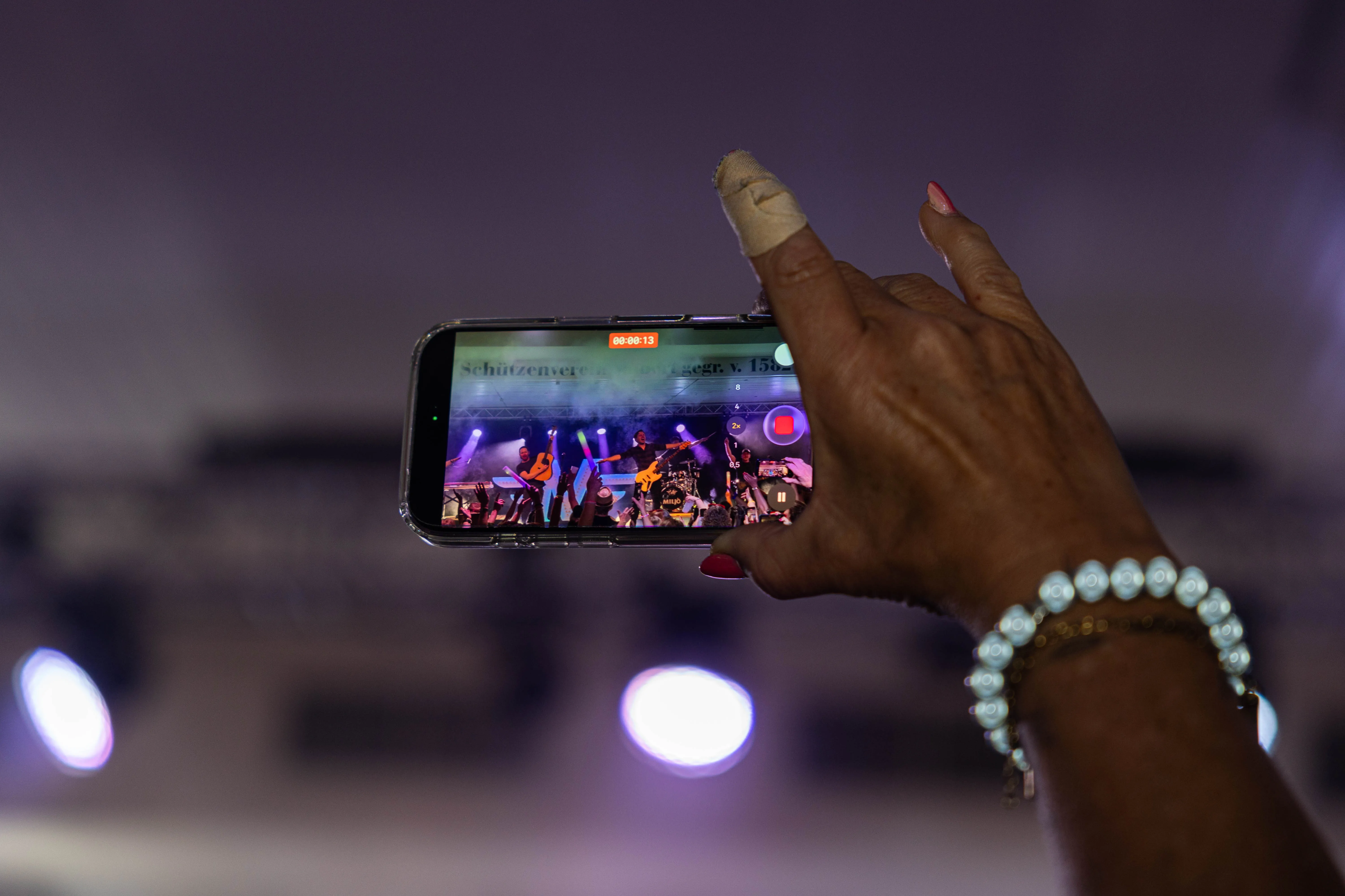 Eine Hand mit rotem Nagellack filmt ein Konzert auf einem Smartphone. Auf dem Bildschirm sind Musiker mit Instrumenten zu sehen.