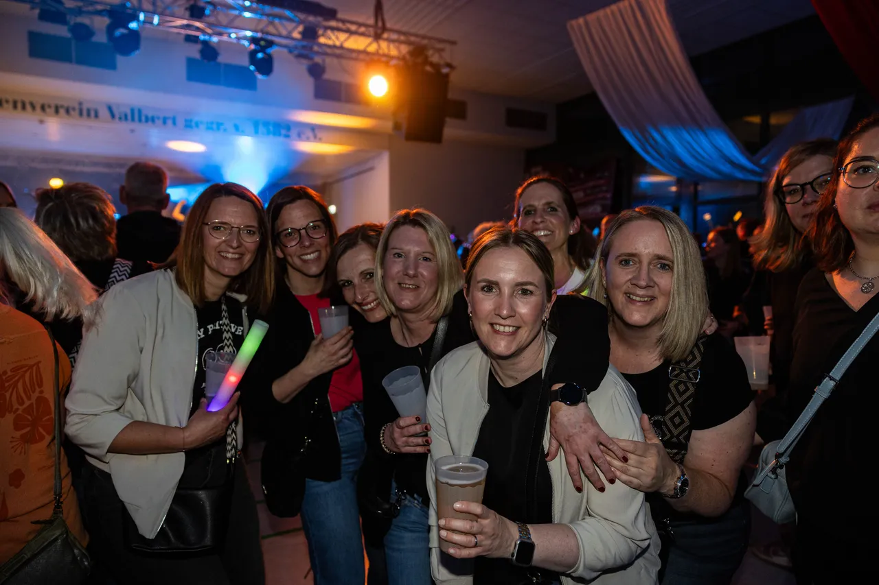 Mehrere lächelnde Frauen stehen eng zusammen, einige halten Getränke in der Hand, im Hintergrund eine festlich beleuchtete Halle.