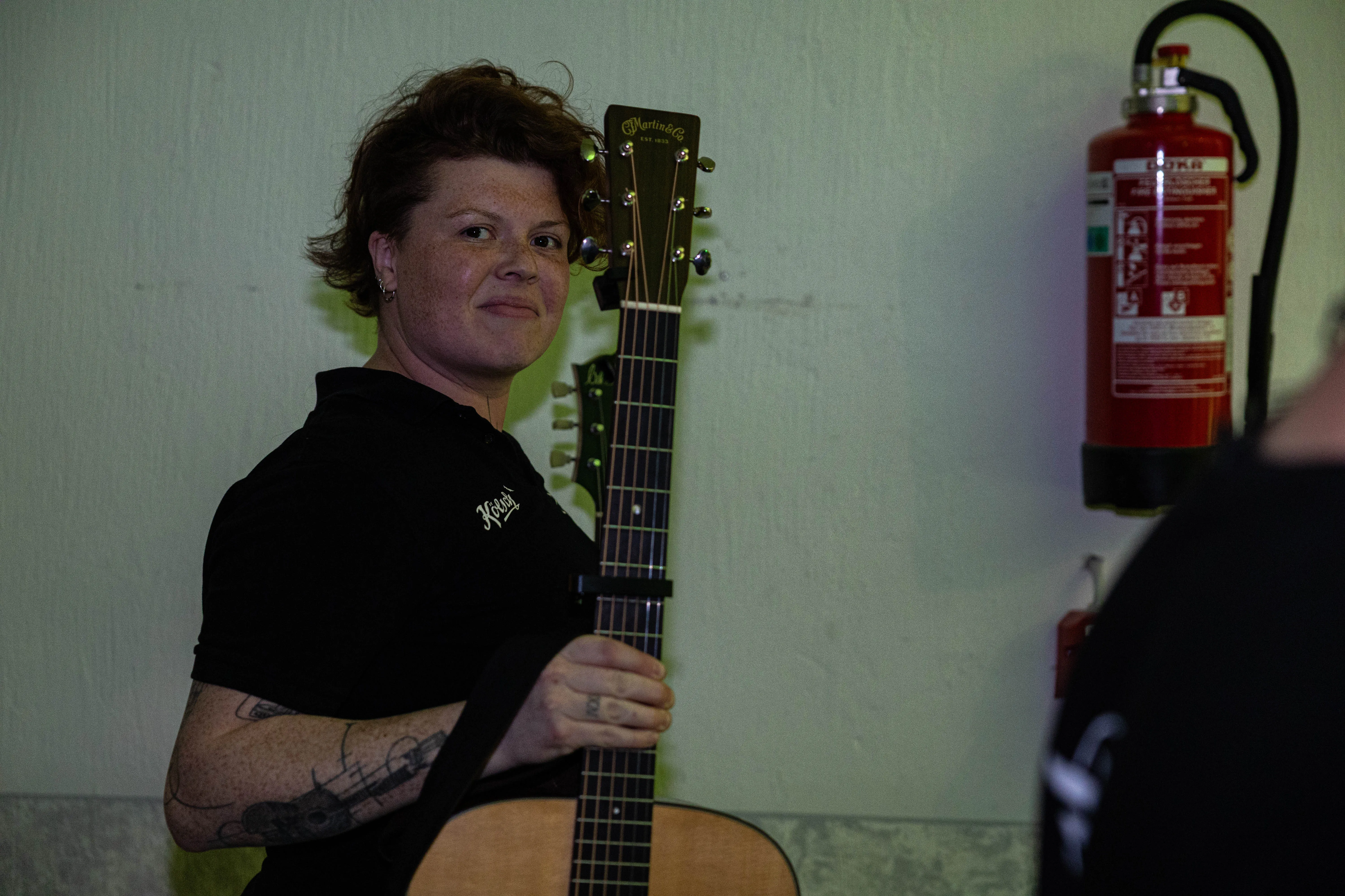 Eine Frau mit roten, lockigen Haaren spielt Gitarre. Im Hintergrund ist ein roter Feuerlöscher an der Wand zu sehen.