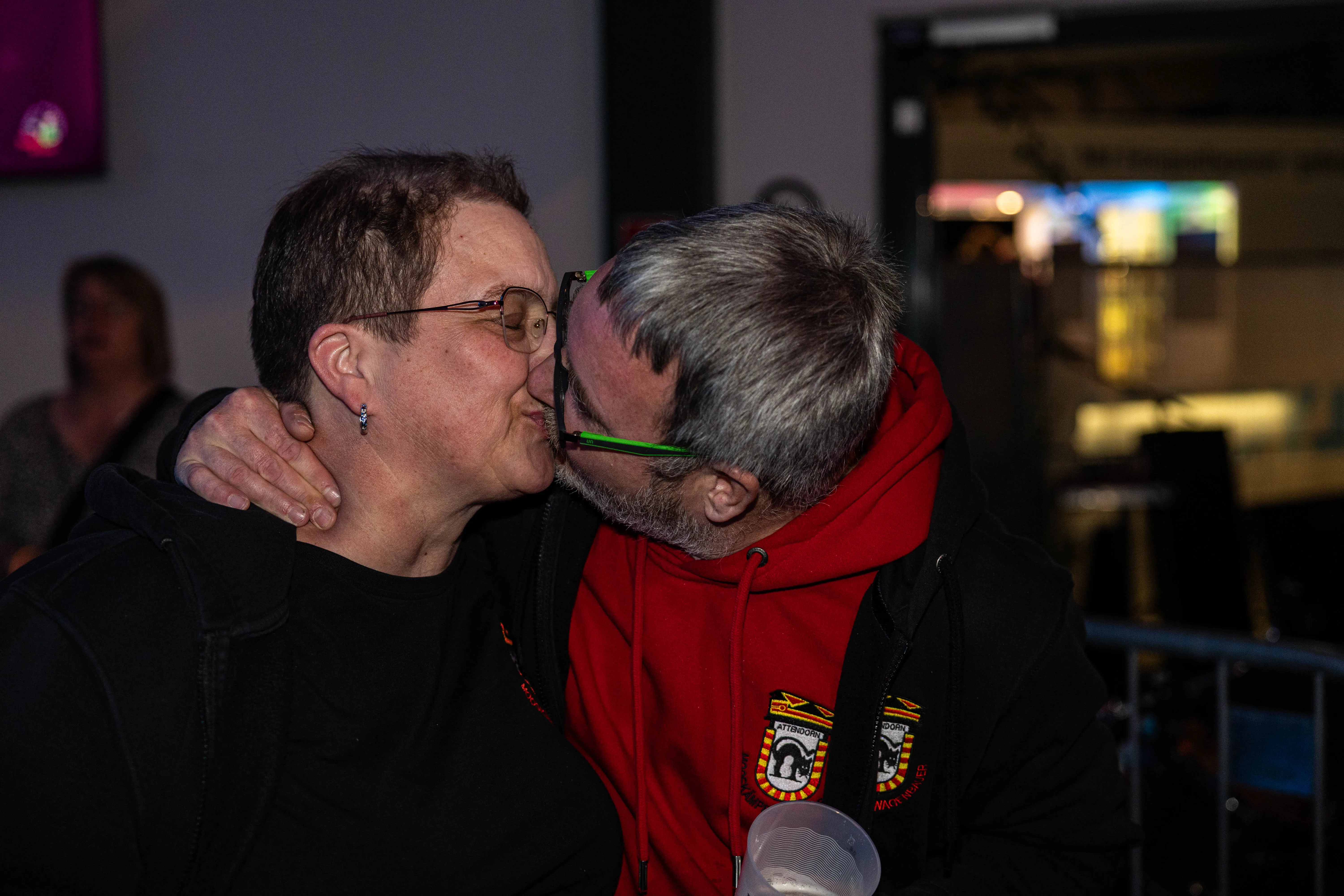 Zwei Personen umarmen sich eng und küssen sich auf der Wange während eines Festivals. Im Hintergrund sind weitere Menschen und eine Bar zu sehen.
