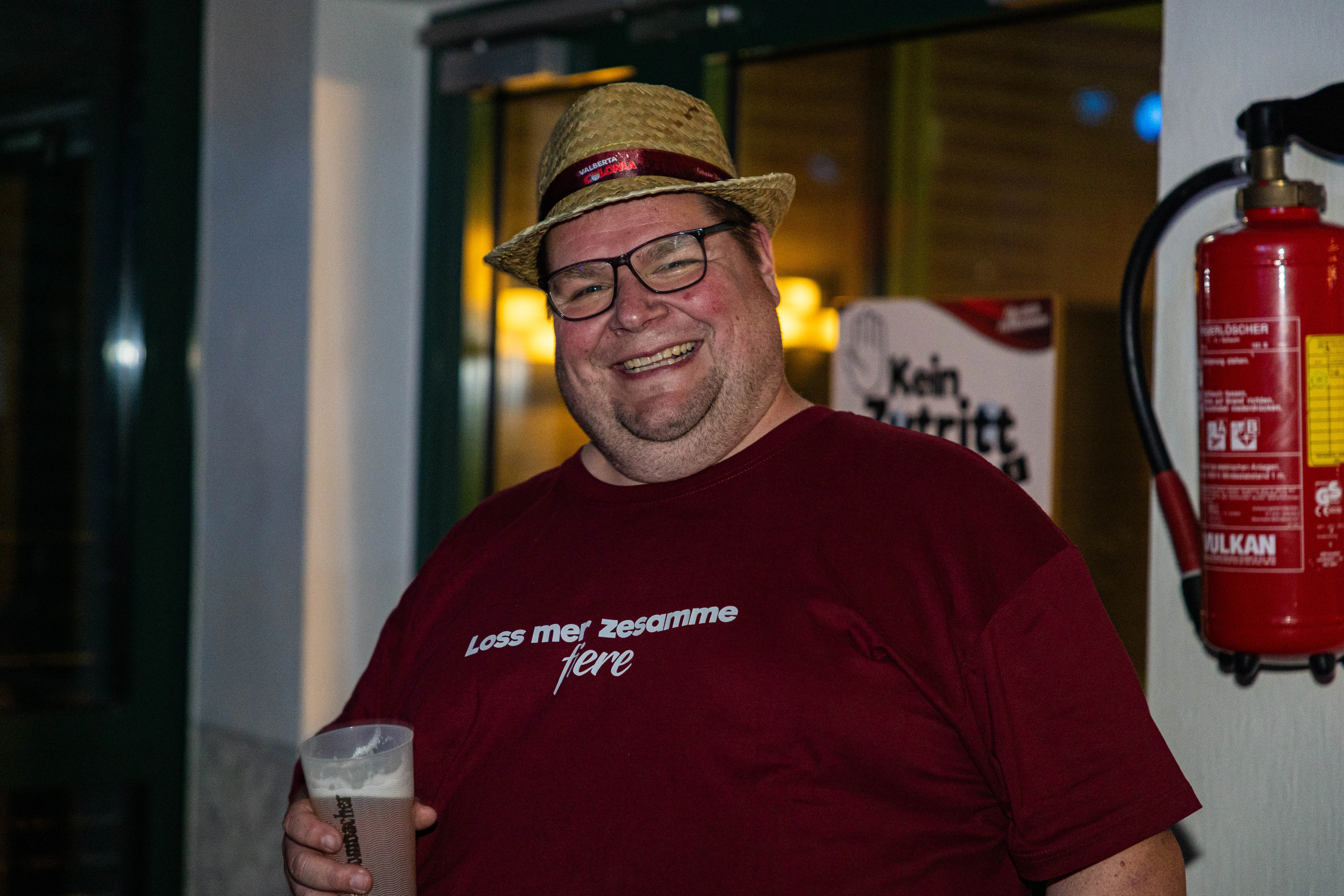 Ein lächelnder Mann mit Strohhut und rotem T-Shirt mit Aufschrift hält ein Bierglas in der Hand. Im Hintergrund ein Schild mit der Aufschrift „Kein Zutritt“ und ein Feuerlöscher.