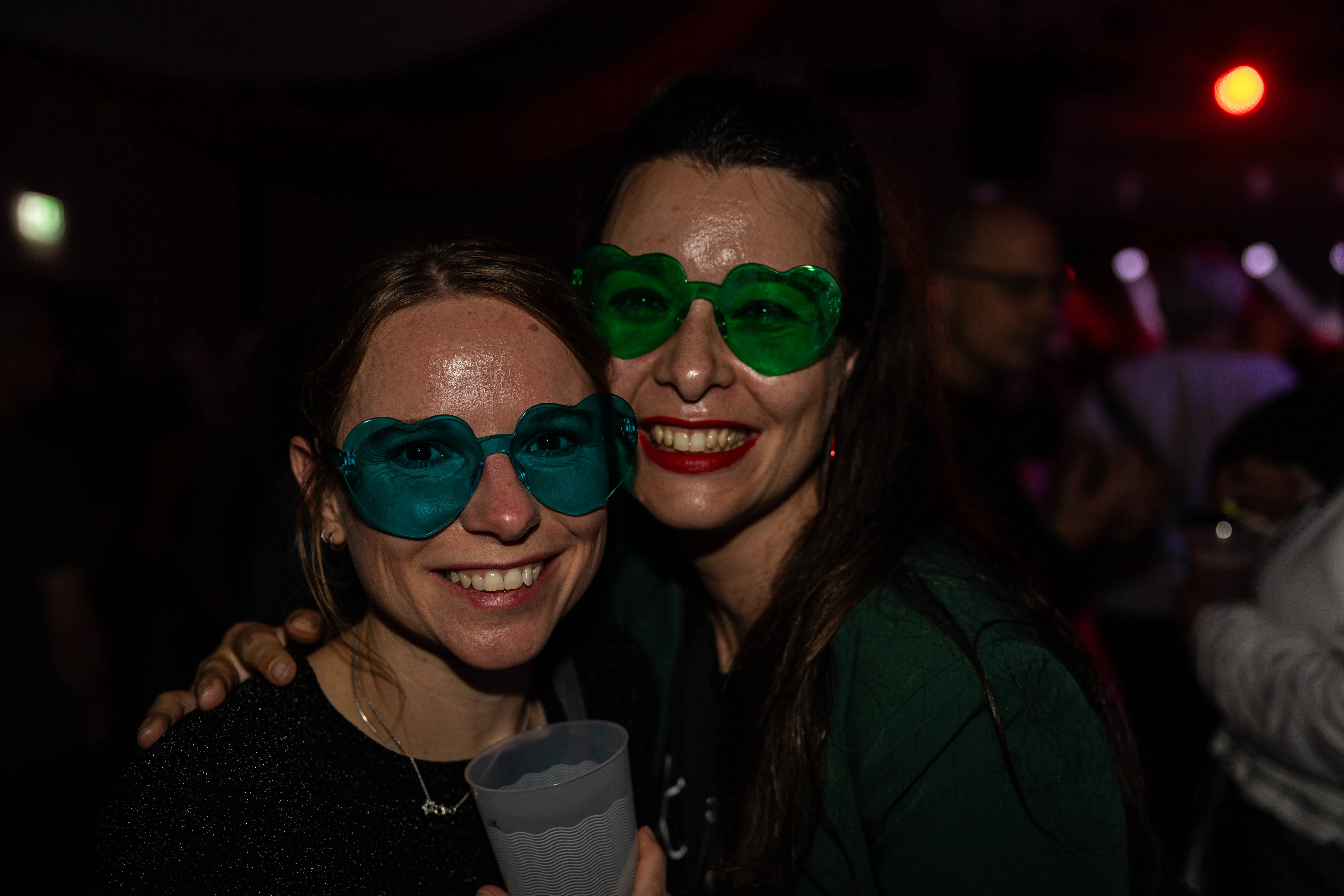 Zwei lächelnde Frauen mit herzförmigen Brillen vor einer verschwommenen Menschenmenge auf dem Kölschmusikfestival Valberta Colonia.