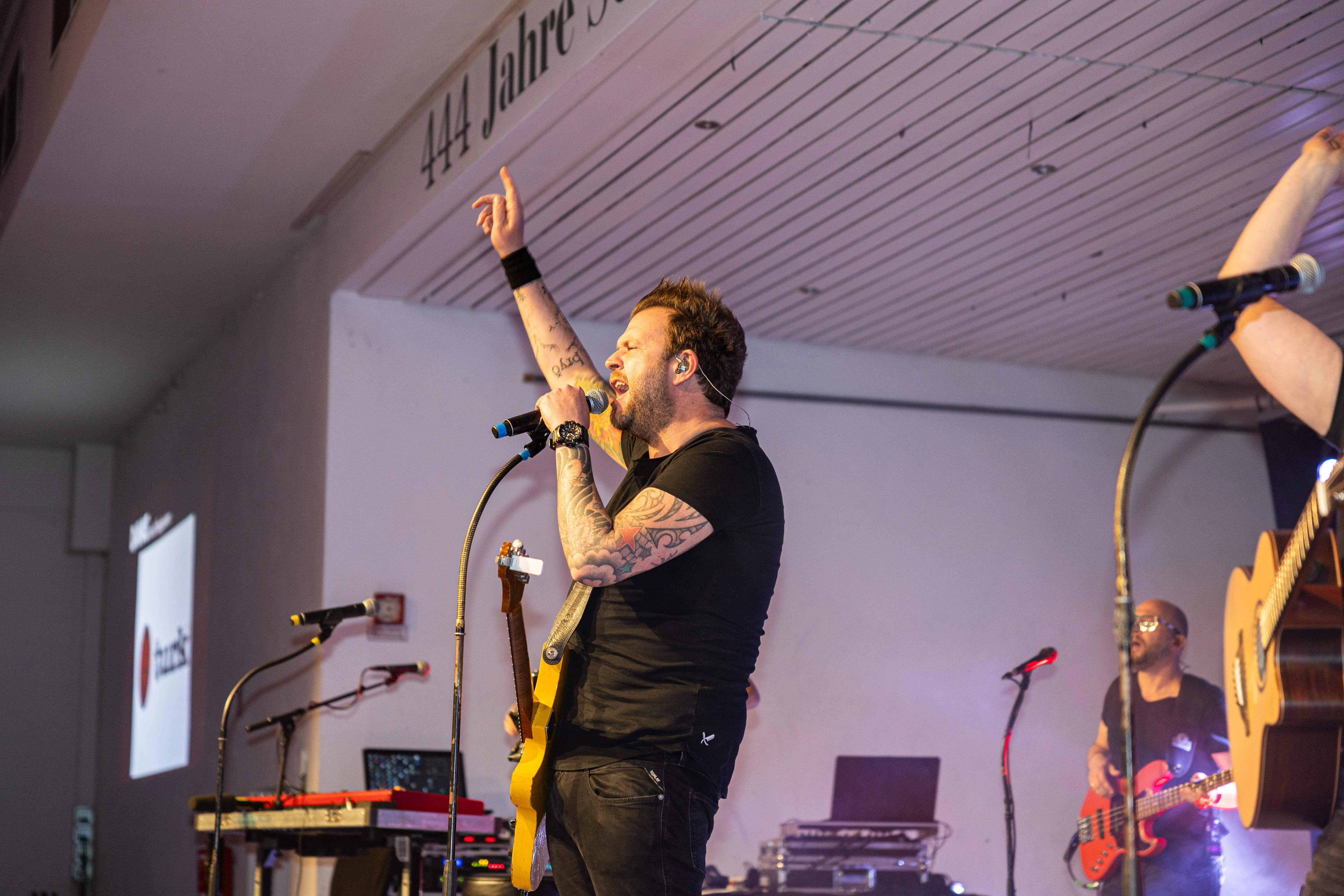 Sänger mit In-Ear-Monitoring und Tätowierungen singt in ein Handmikrofon, während er eine Gitarre hält und die Hand hebt. Im Hintergrund ein weiterer Musiker mit Gitarre auf einer Bühne.