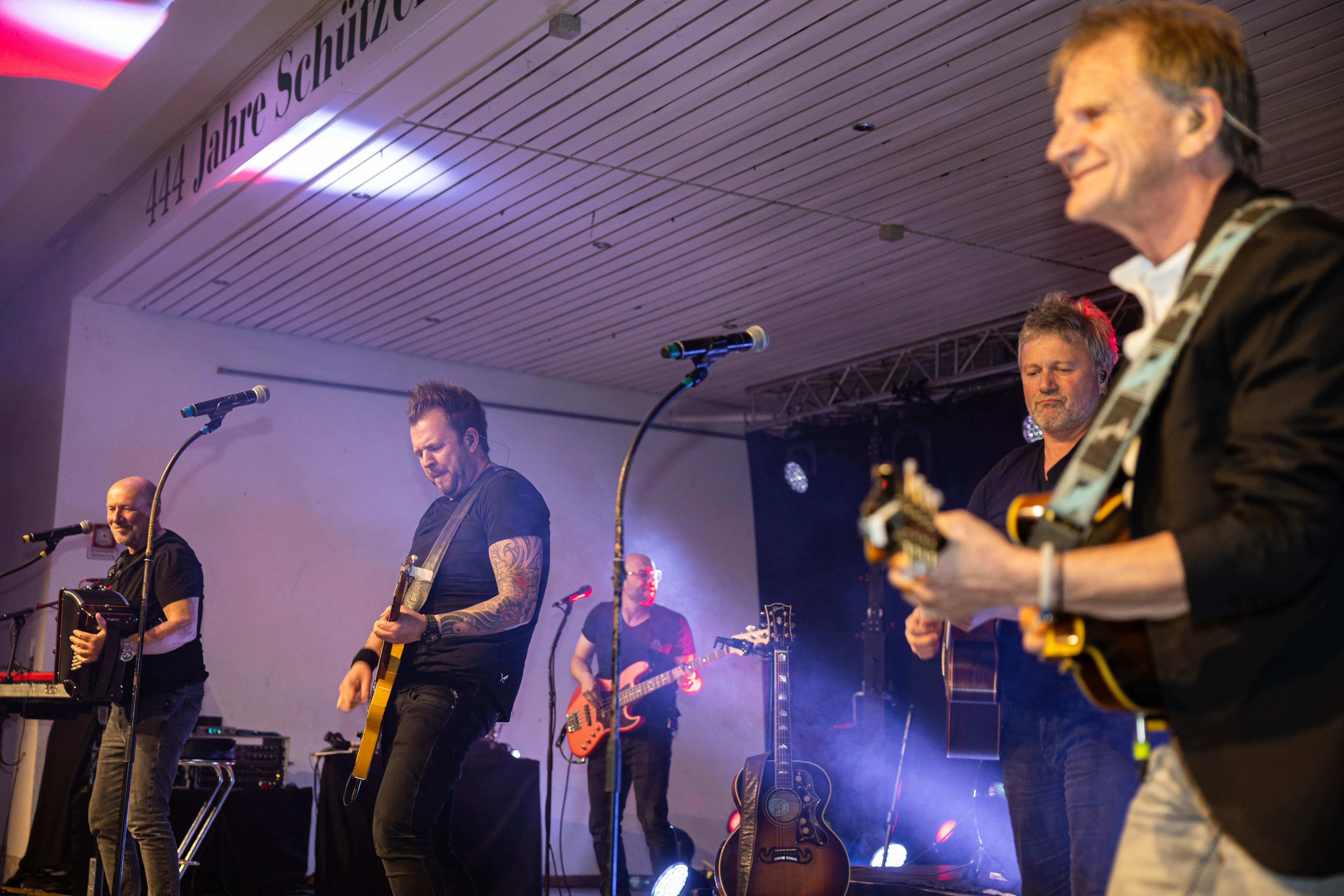 Vier Musiker spielen auf einer Bühne vor Publikum. Im Hintergrund ist ein Banner mit der Aufschrift „444 Jahre Schützenverein“ zu sehen.