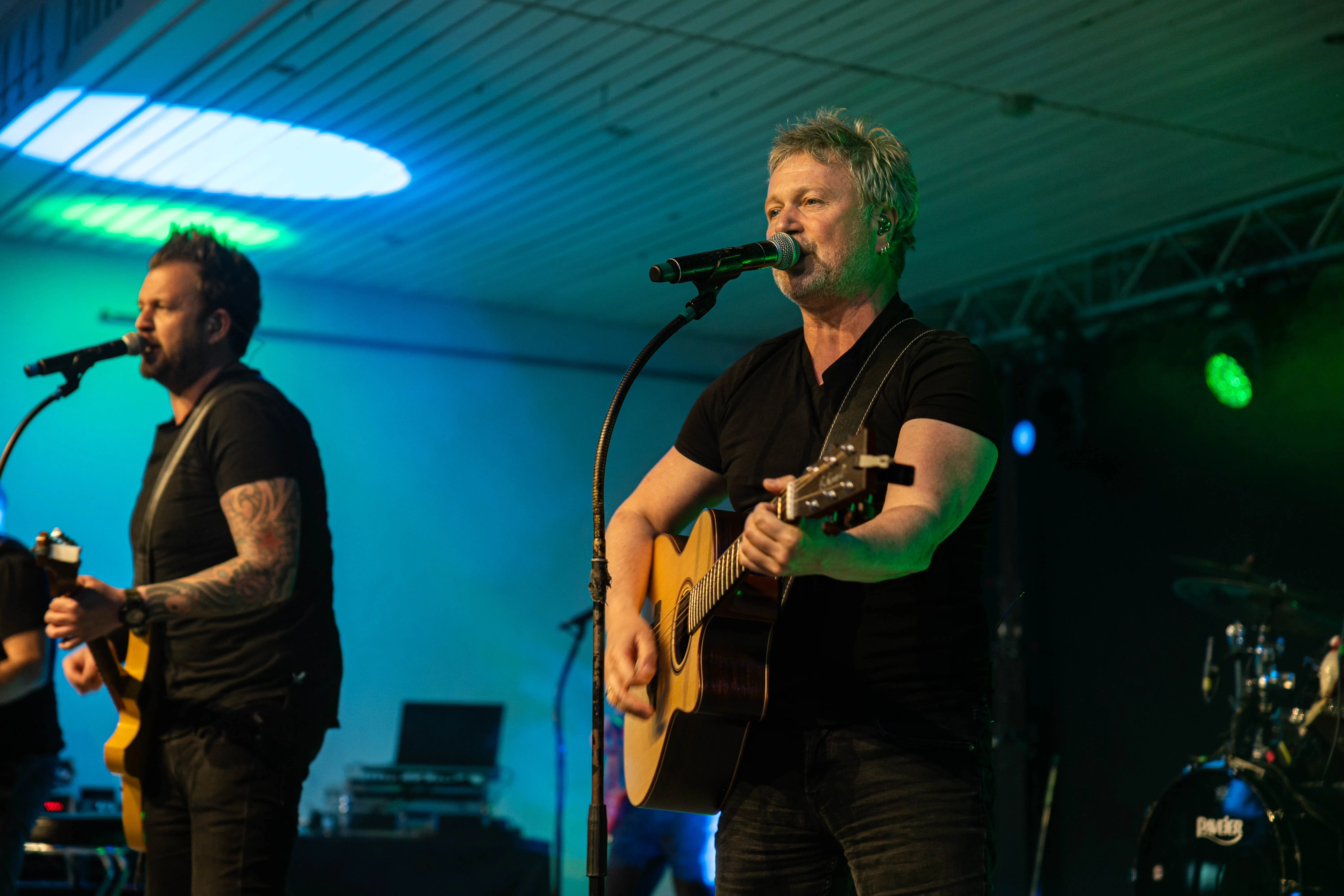 Zwei Musiker auf einer Bühne, der eine spielt Gitarre und singt ins Mikrofon, der andere singt ebenfalls ins Mikrofon und hält eine Gitarre. Im Hintergrund ist eine blaue Beleuchtung und ein Schlagzeug zu sehen.