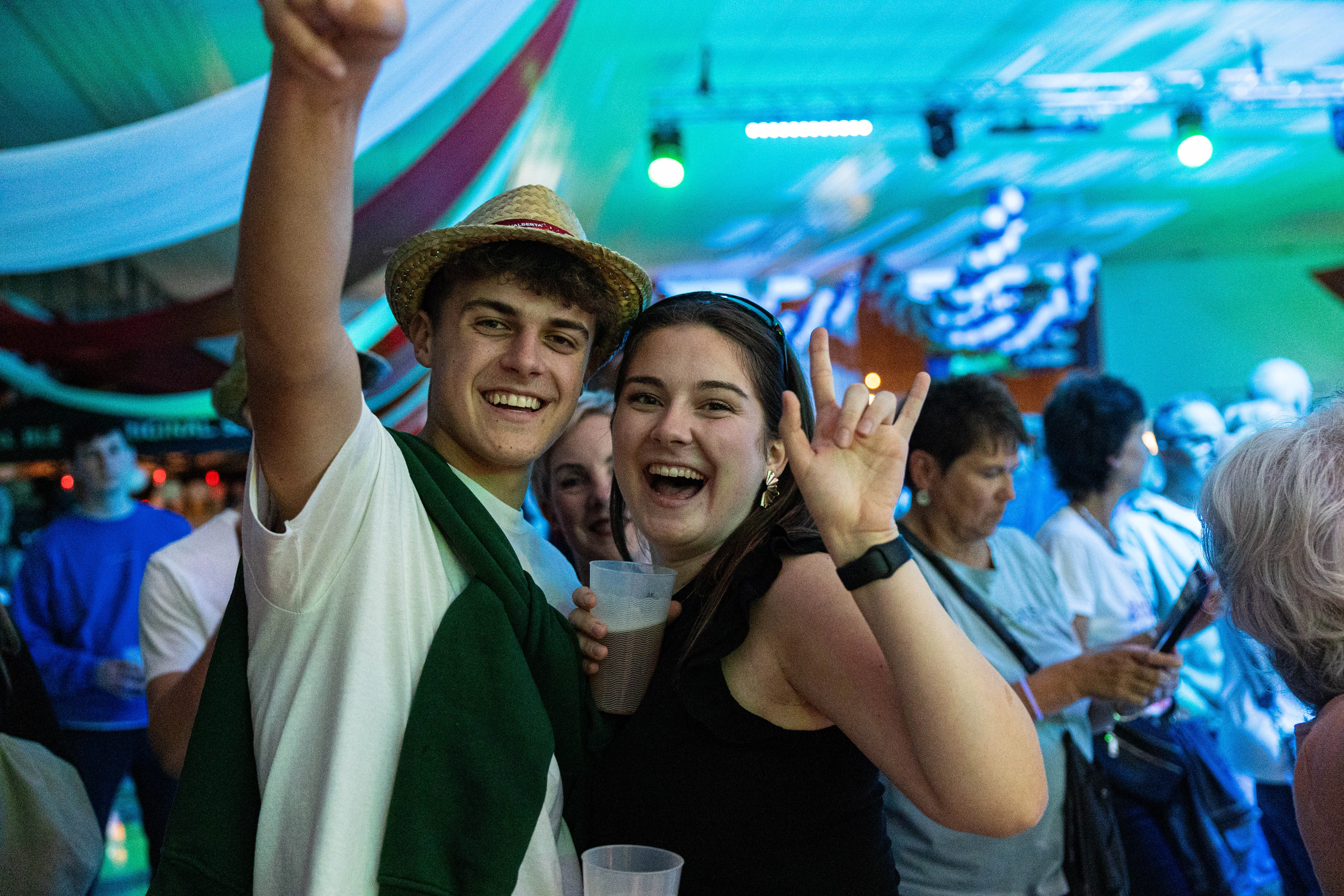 Zwei junge Menschen lächeln in die Kamera, der Mann mit hochgehobenem Arm, die Frau mit Handzeichen. Im Hintergrund eine Menschenmenge auf dem Kölschmusikfestival Valberta Colonia.