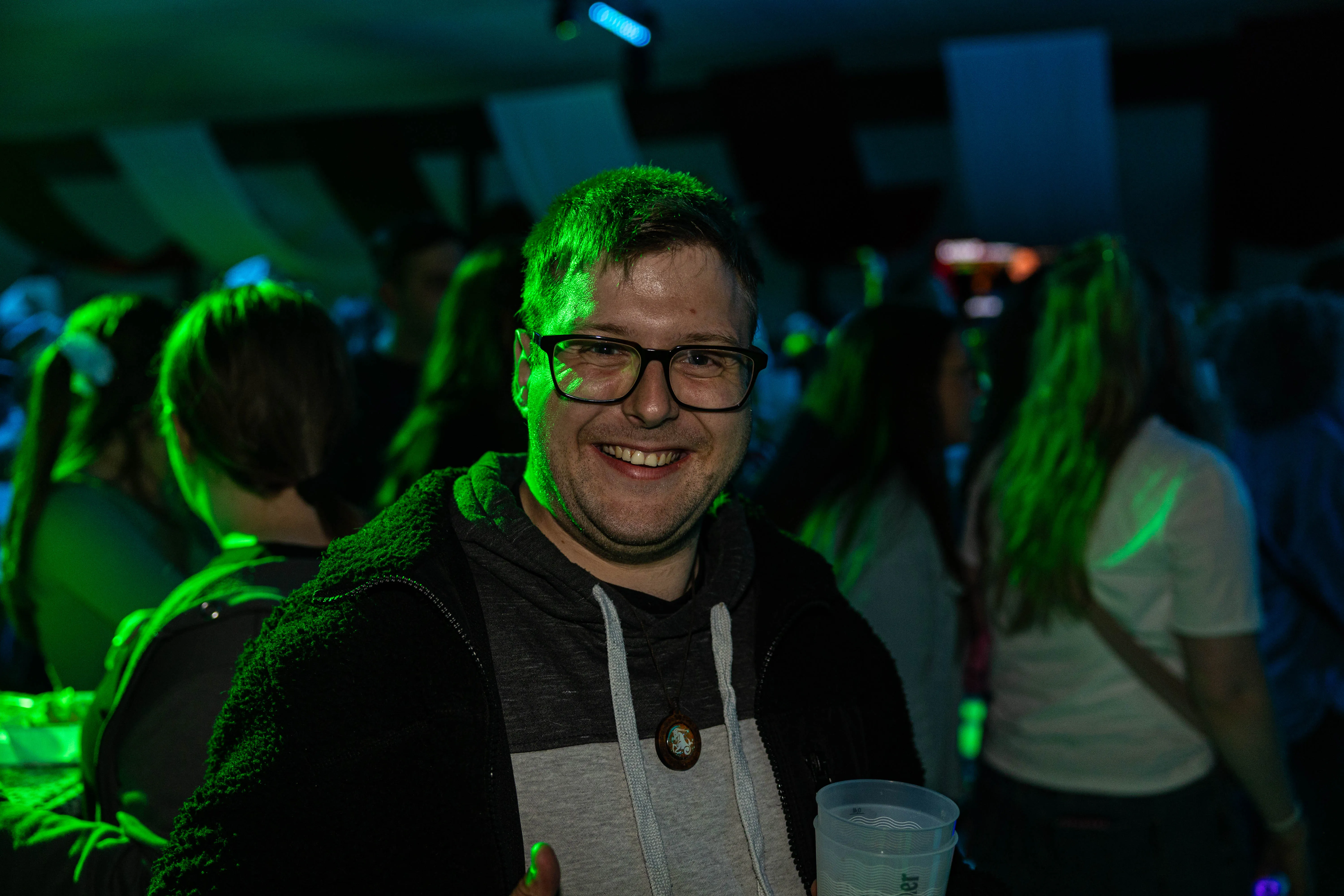 Lachender Mann mit Brille und Hoodie hält ein Getränk in der Hand, im Hintergrund Menschenmenge bei einem Konzert.