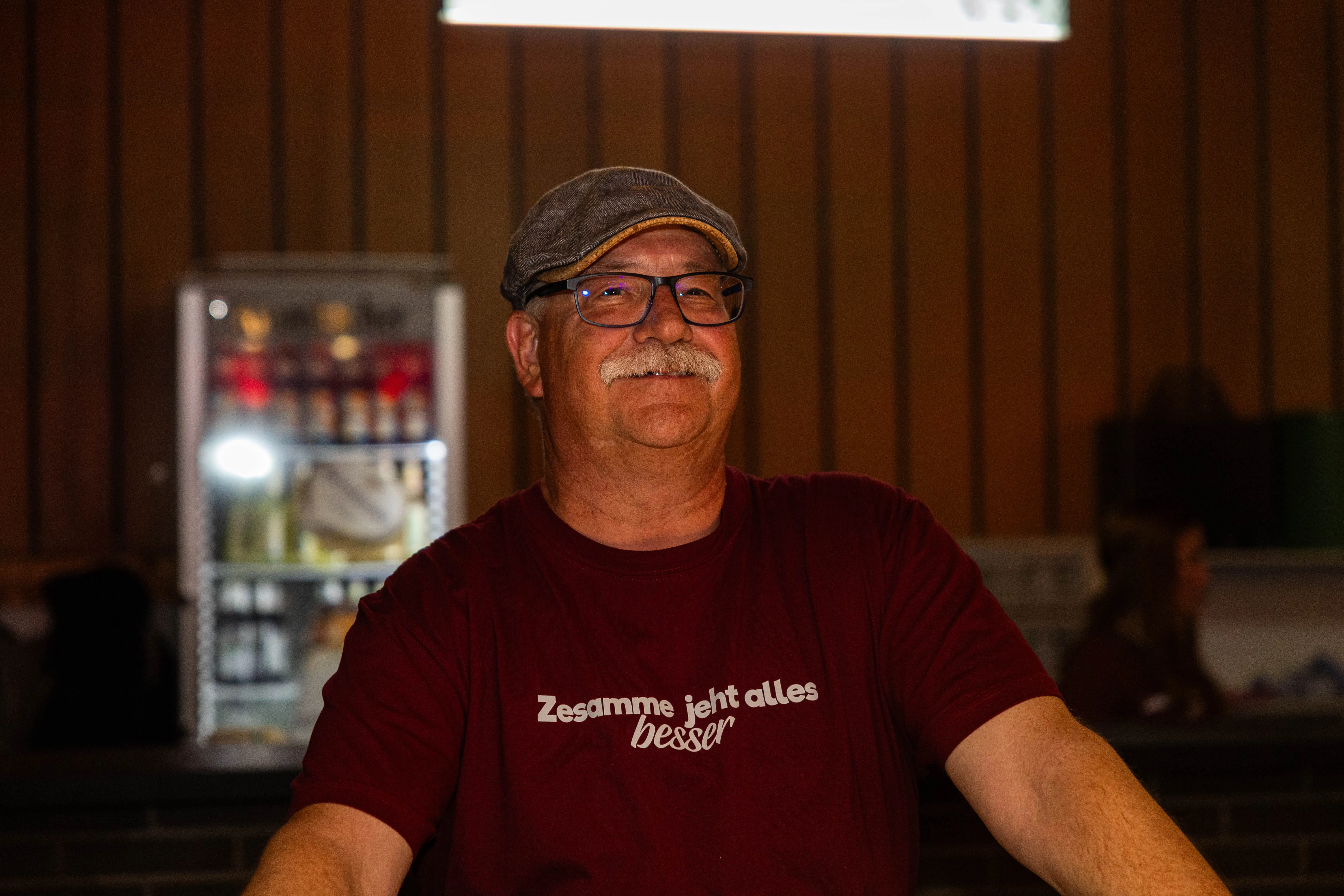 Mann mit Schnurrbart und Brille trägt eine Kappe und ein rotes T-Shirt mit Aufschrift, lächelt in die Kamera. Im Hintergrund ein Getränkekühlschrank und verschwommene Personen.