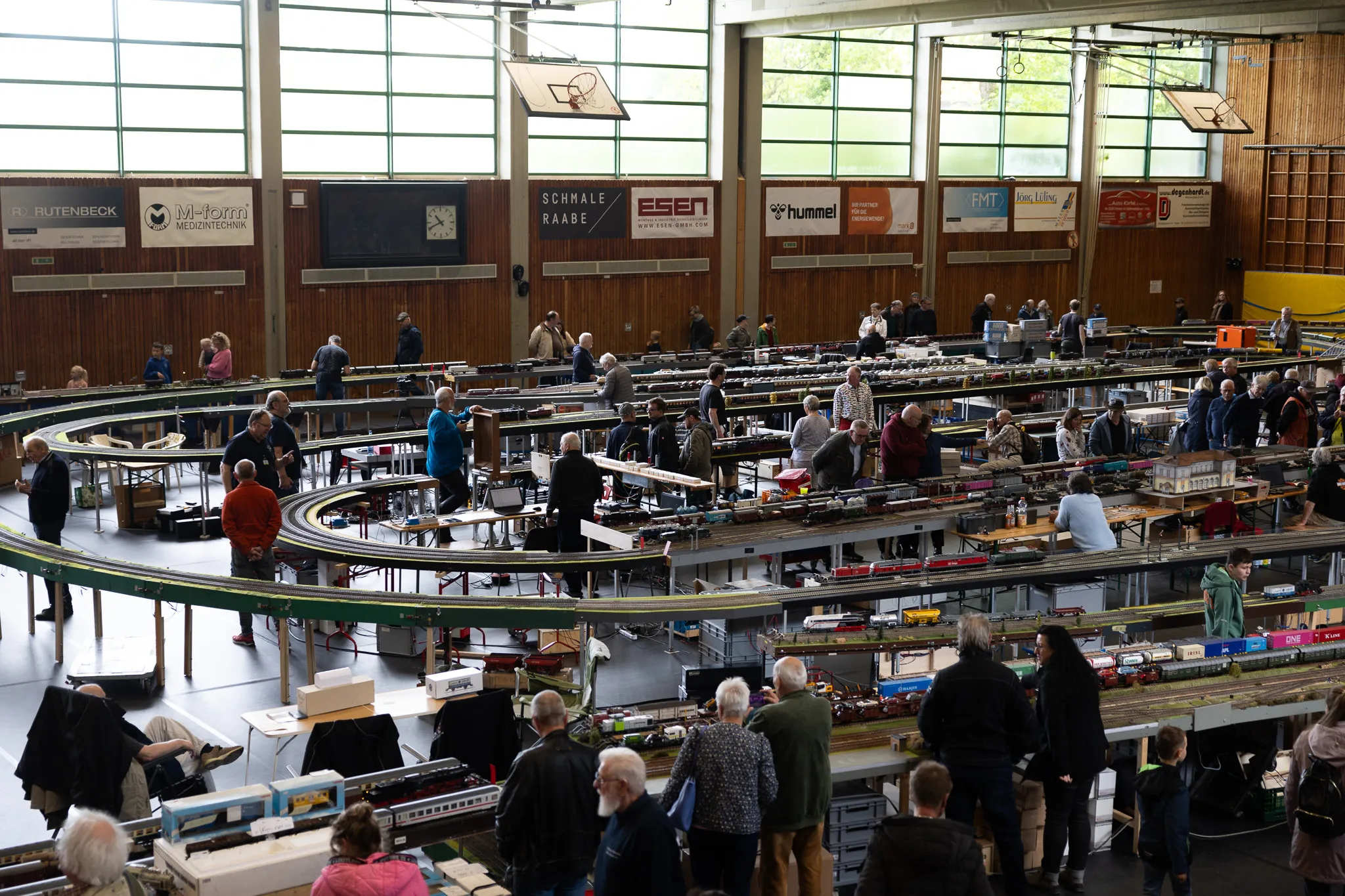 Große Modellbahn-Ausstellung in einer Turnhalle mit vielen Besuchern, die die Anlage umrunden und die Züge beobachten.