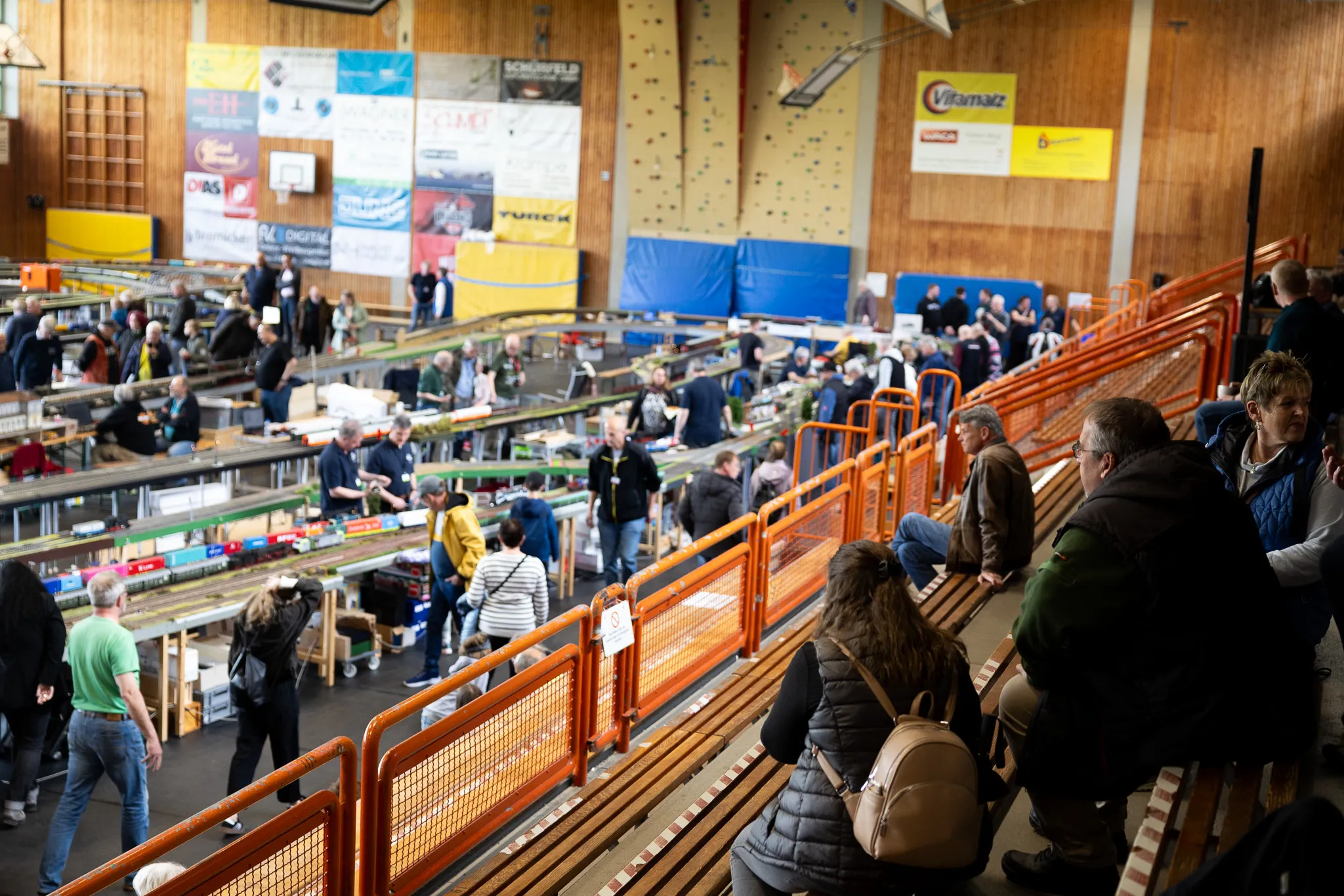 Innenansicht einer gut besuchten Modellbahn-Ausstellung in einer Turnhalle mit zahlreichen Besuchern vor langen Tischen voller Modelleisenbahnen.