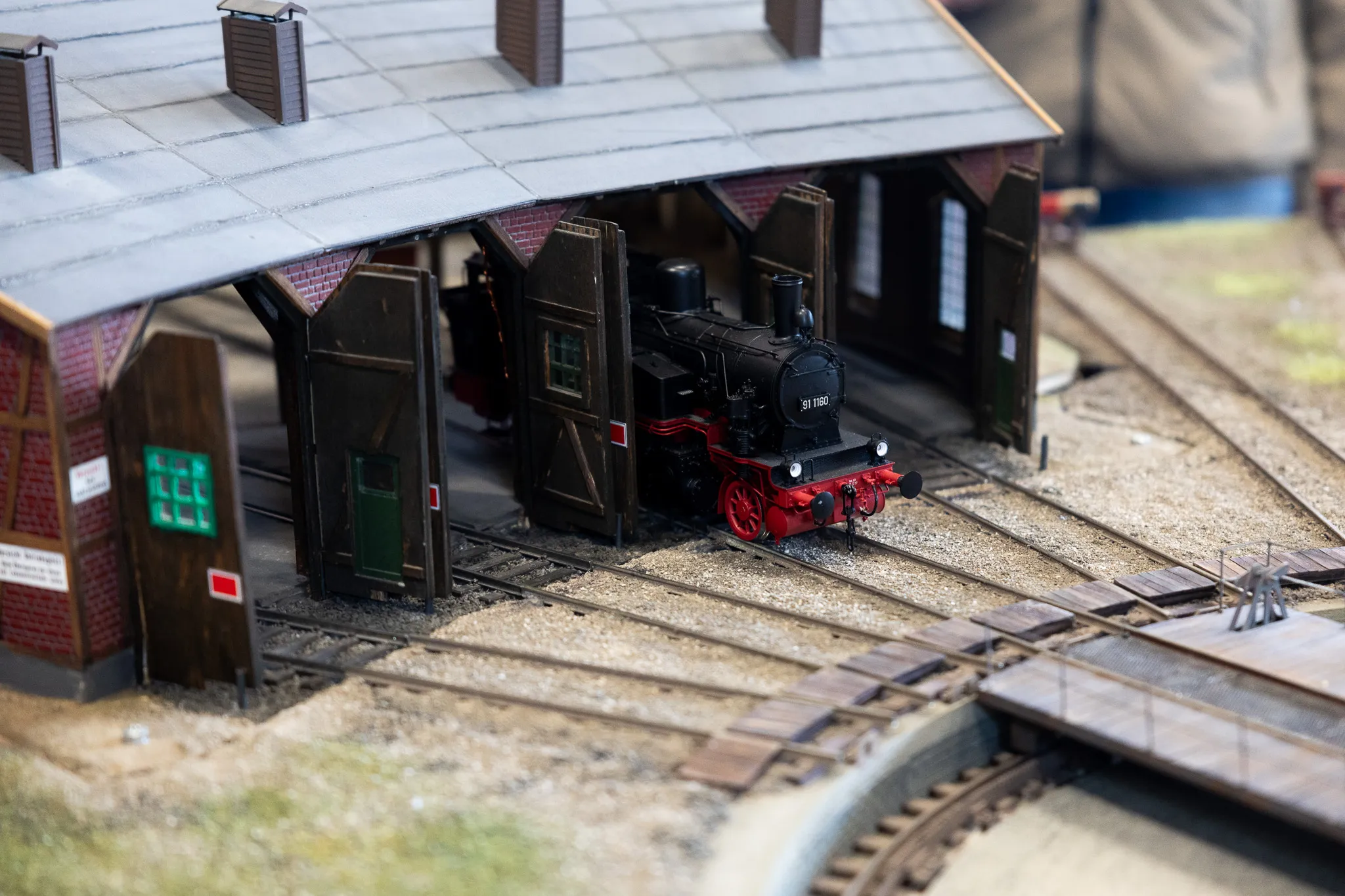 Eine Dampflokomotive steht in einem Modellbahnhof unter einem hölzernen Dach. Gleise kreuzen sich vor dem Bahnhofsgebäude.